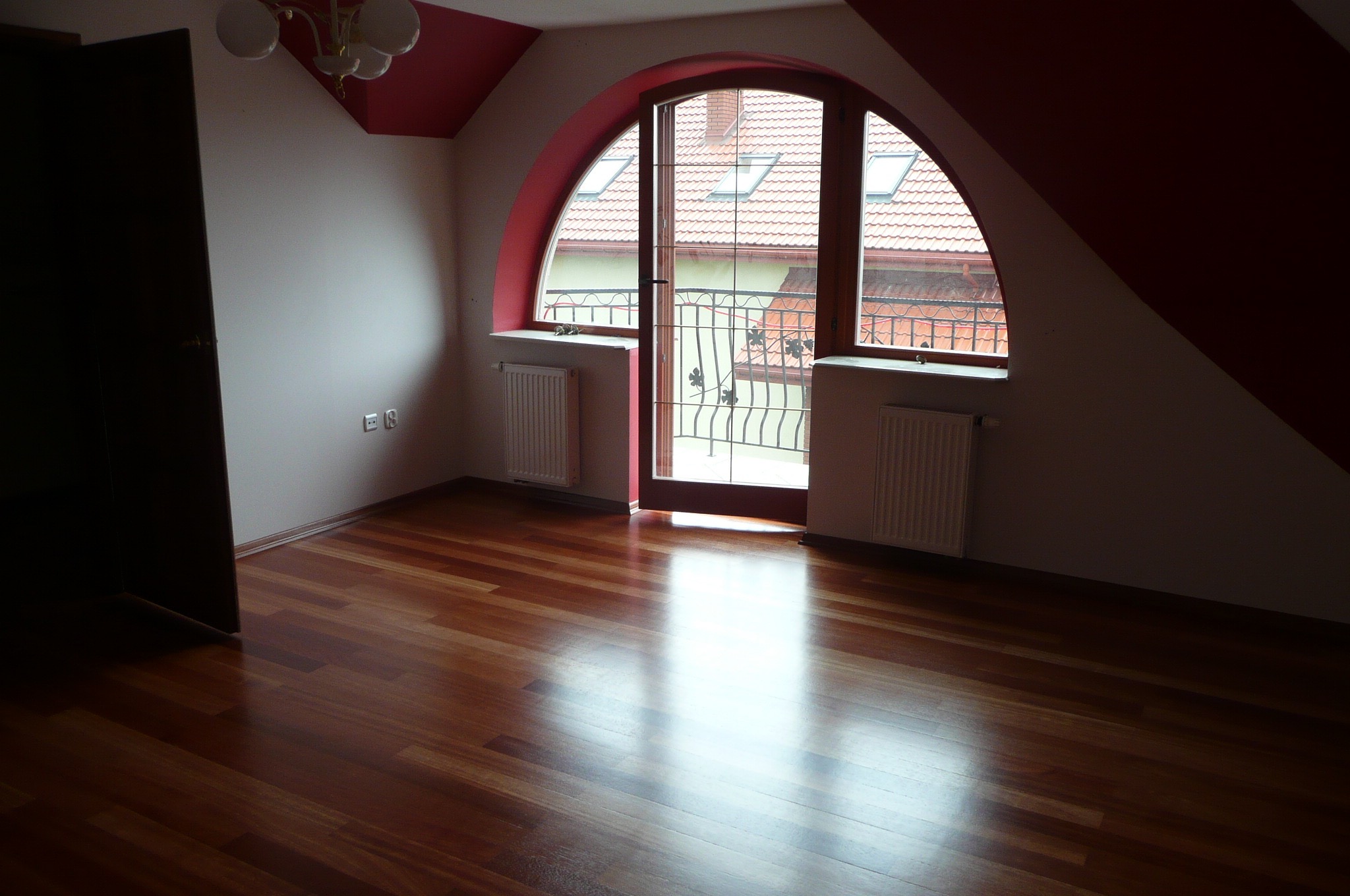 Pokój na poddaszu z wypolerowaną, drewnianą podłogą, owalnym oknem z widokiem na dach sąsiedniego budynku i grzejnikiem pod oknem.