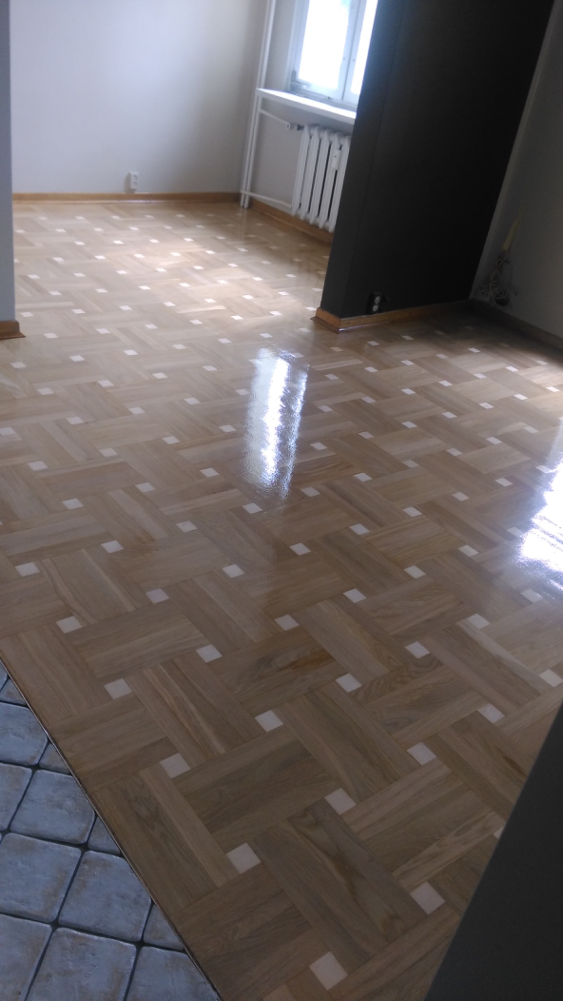 Parkiet ułożony w jodełkę po renowacji, z widocznymi białymi wstawkami, błyszczący od świeżego lakieru, z fragmentem płytek podłogowych w rogu pokoju.