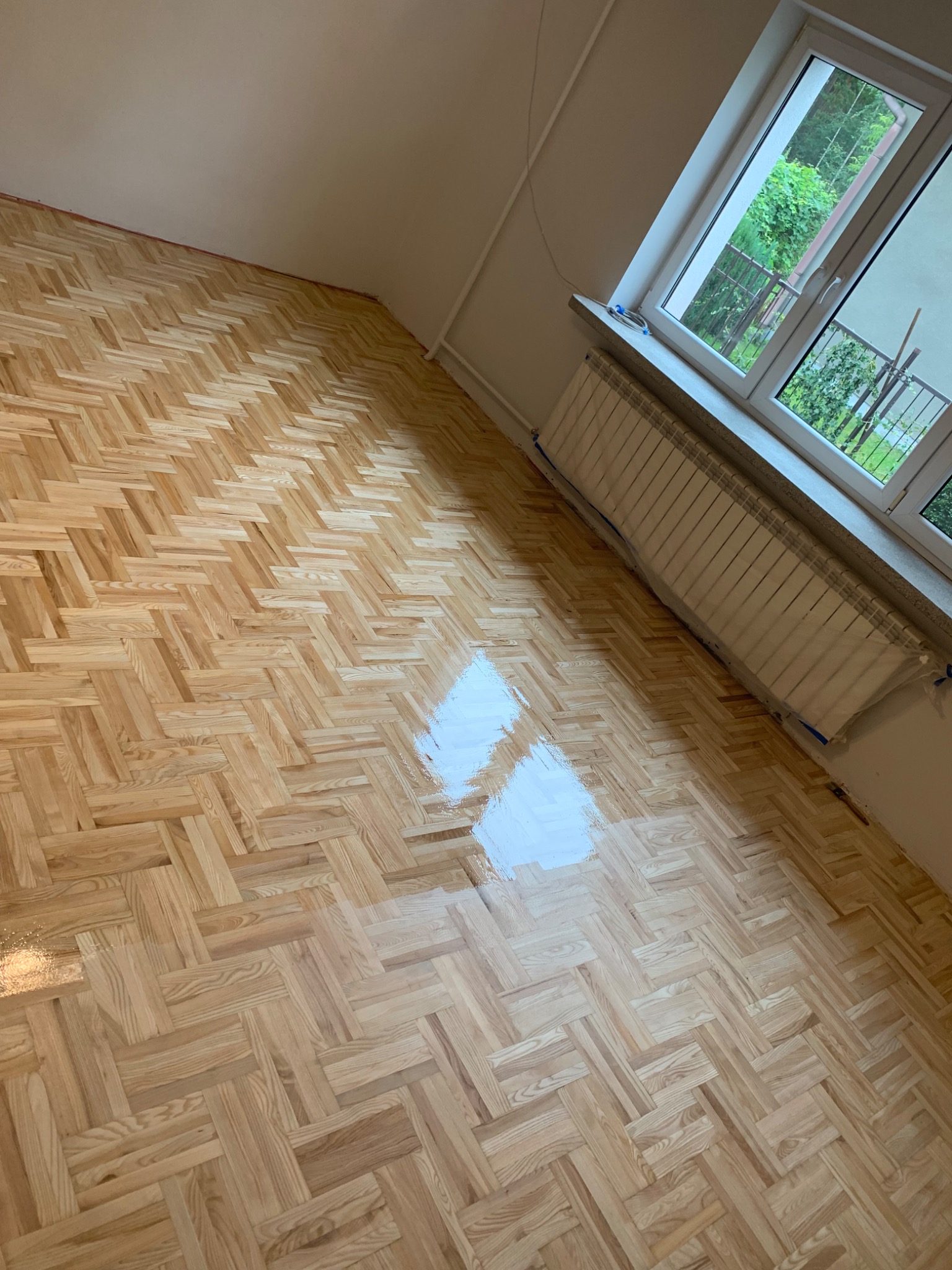 Parkiet ułożony w jodełkę, częściowo pokryty świeżym lakierem, odbijającym światło z okna. Grzejnik pod oknem zabezpieczony folią malarską.