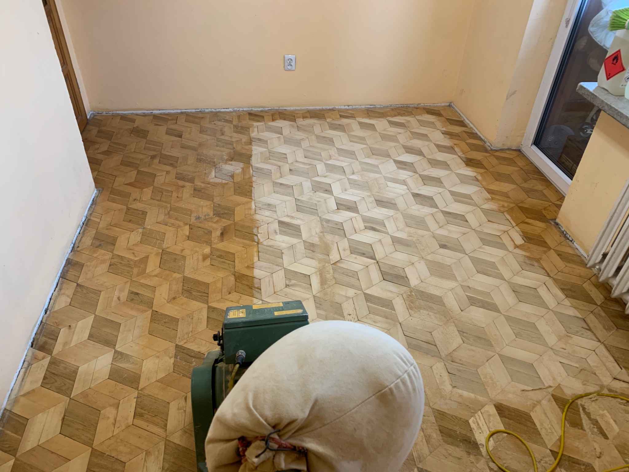 Parkiet w geometryczny wzór, częściowo odnowiony maszyną do cyklinowania, widoczny worek na pył i kabel zasilający.