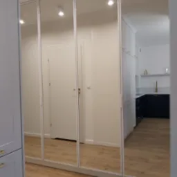 Szafa z lustrzanymi drzwiami przesuwnymi, odbijająca wnętrze mieszkania z widoczną kuchnią w tle, jasne drewniane podłogi.