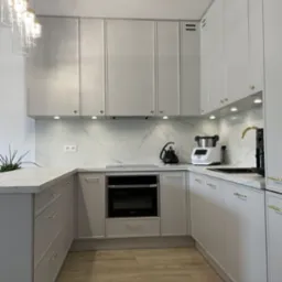 Jasnoszara kuchnia z marmurowym blatem i backsplashiem, złote uchwyty, nowoczesne oświetlenie podszafkowe, czajnik i robot kuchenny na blacie, drewniana podłoga.