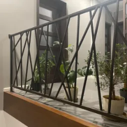 Wewnętrzna balustrada z metalu o geometrycznym wzorze, z roślinami doniczkowymi na tle szarej podłogi i ciemnych drzwi.