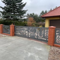 Augustowiak - Metalowa brama wjazdowa i przęsła ogrodzenia z motywem wycinanych laserowo sylwetek drzew, ptaków i roślinności wodnej, osadzone na słupkach z czerwonej cegły, na tle domu z czerwoną dachówką...