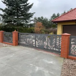 Metalowa brama wjazdowa i przęsła ogrodzenia z motywem wycinanych laserowo sylwetek drzew, ptaków i roślinności wodnej, osadzone na słupkach z czerwonej cegły, na tle domu z czerwoną dachówką...