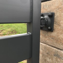 Augustowiak - Szczegół mocowania metalowej bramy wjazdowej do murowanego słupka, widoczne śruby i spawane łączenia, trawa w tle