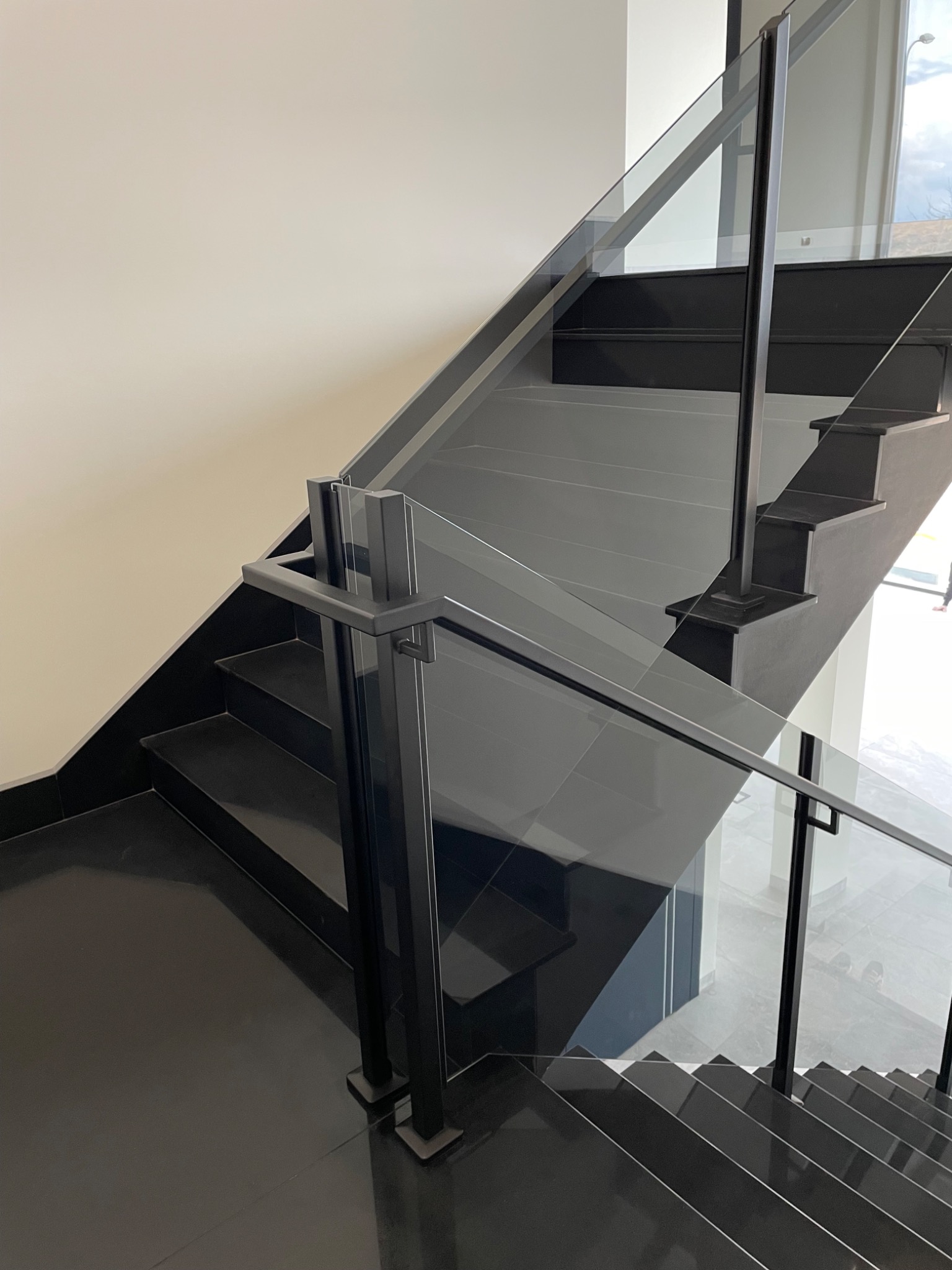 Nowoczesne schody z czarnymi stopniami i szklaną balustradą z czarnymi, metalowymi słupkami.