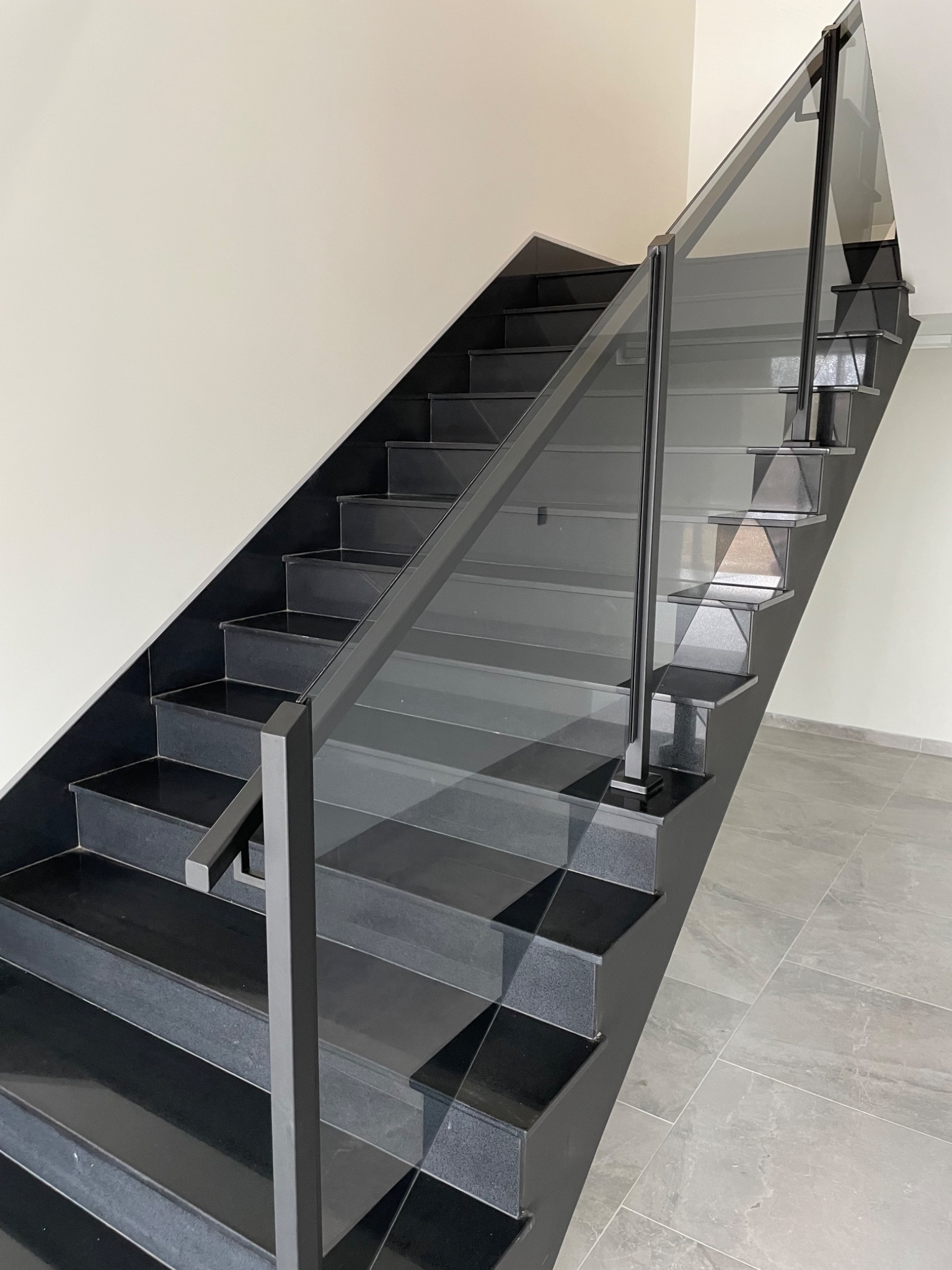 Nowoczesne schody z czarnymi stopniami i szklaną balustradą, widok z dołu, minimalistyczny design, jasne ściany i szare płytki podłogowe.