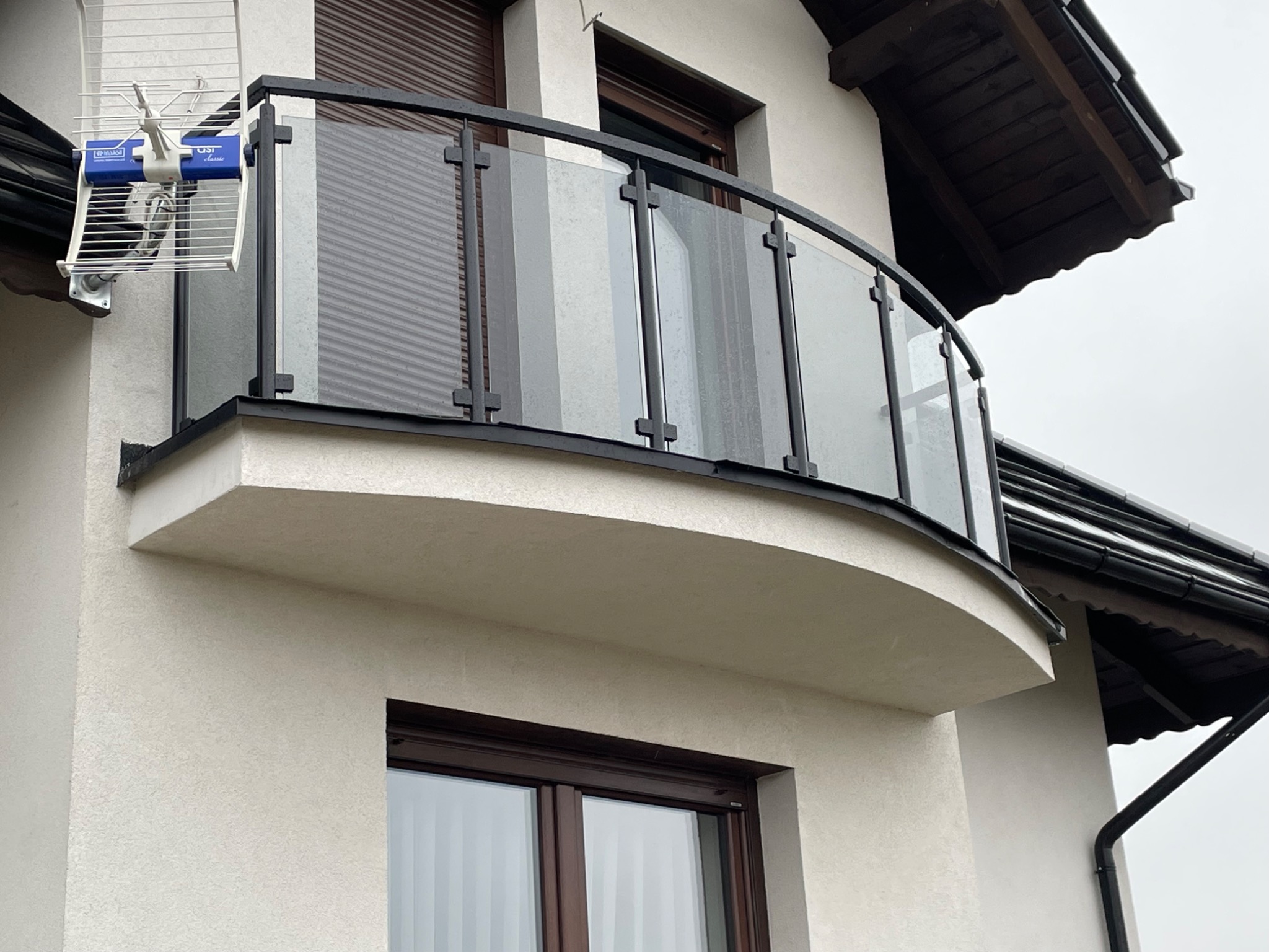 Zaokrąglony balkon z czarną ramą i szklanymi panelami, na tle jasnej elewacji domu i anteny telewizyjnej.