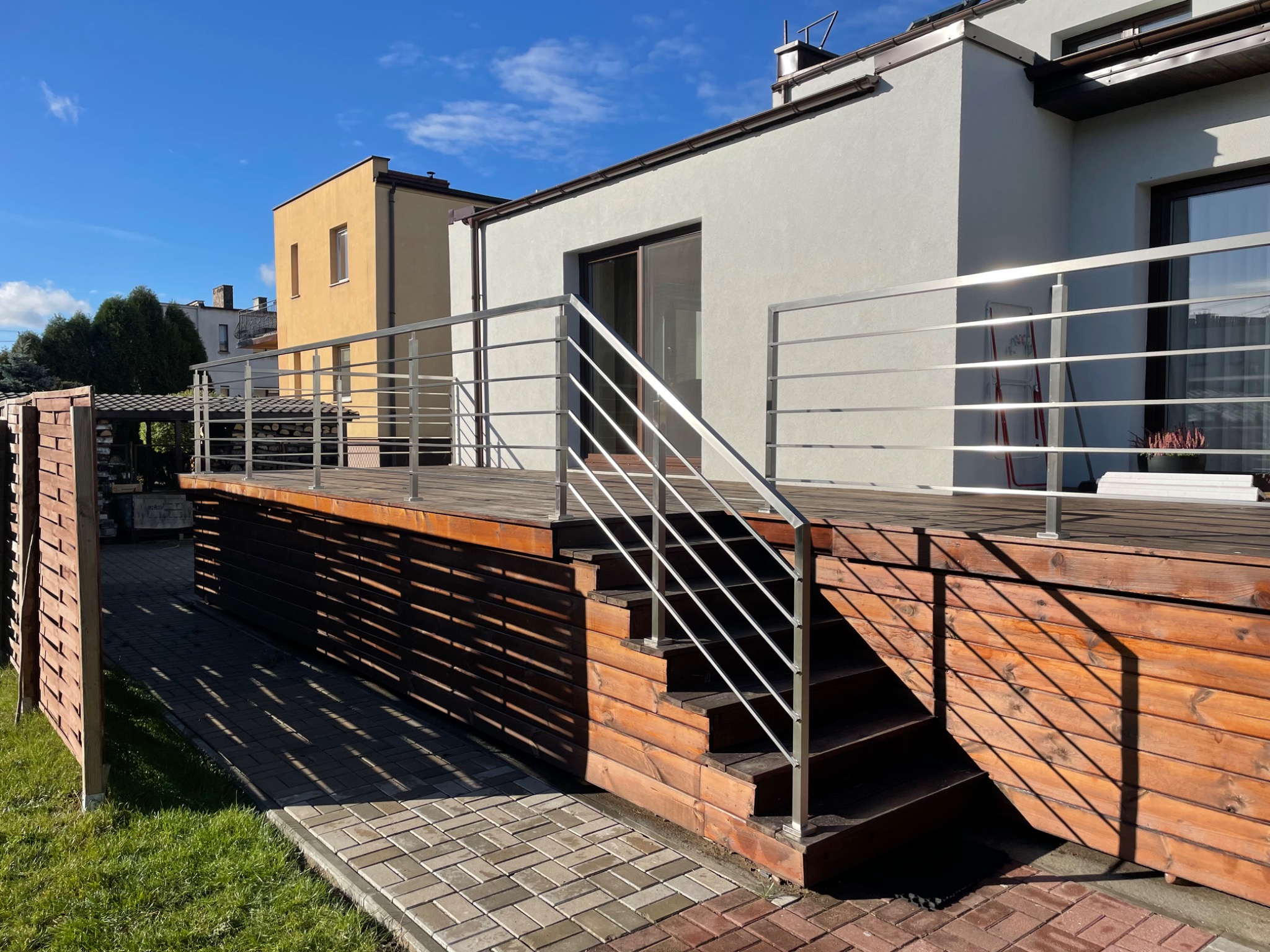 Zewnętrzne schody drewniane z metalową balustradą na tarasie przy nowoczesnym domu w słoneczny dzień.