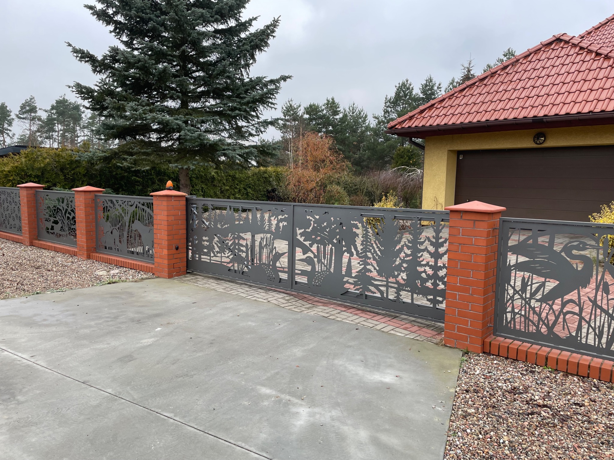 Metalowa brama wjazdowa i przęsła ogrodzenia z motywem wycinanych laserowo sylwetek drzew, ptaków i roślinności wodnej, osadzone na słupkach z czerwonej cegły, na tle domu z czerwoną dachówką...