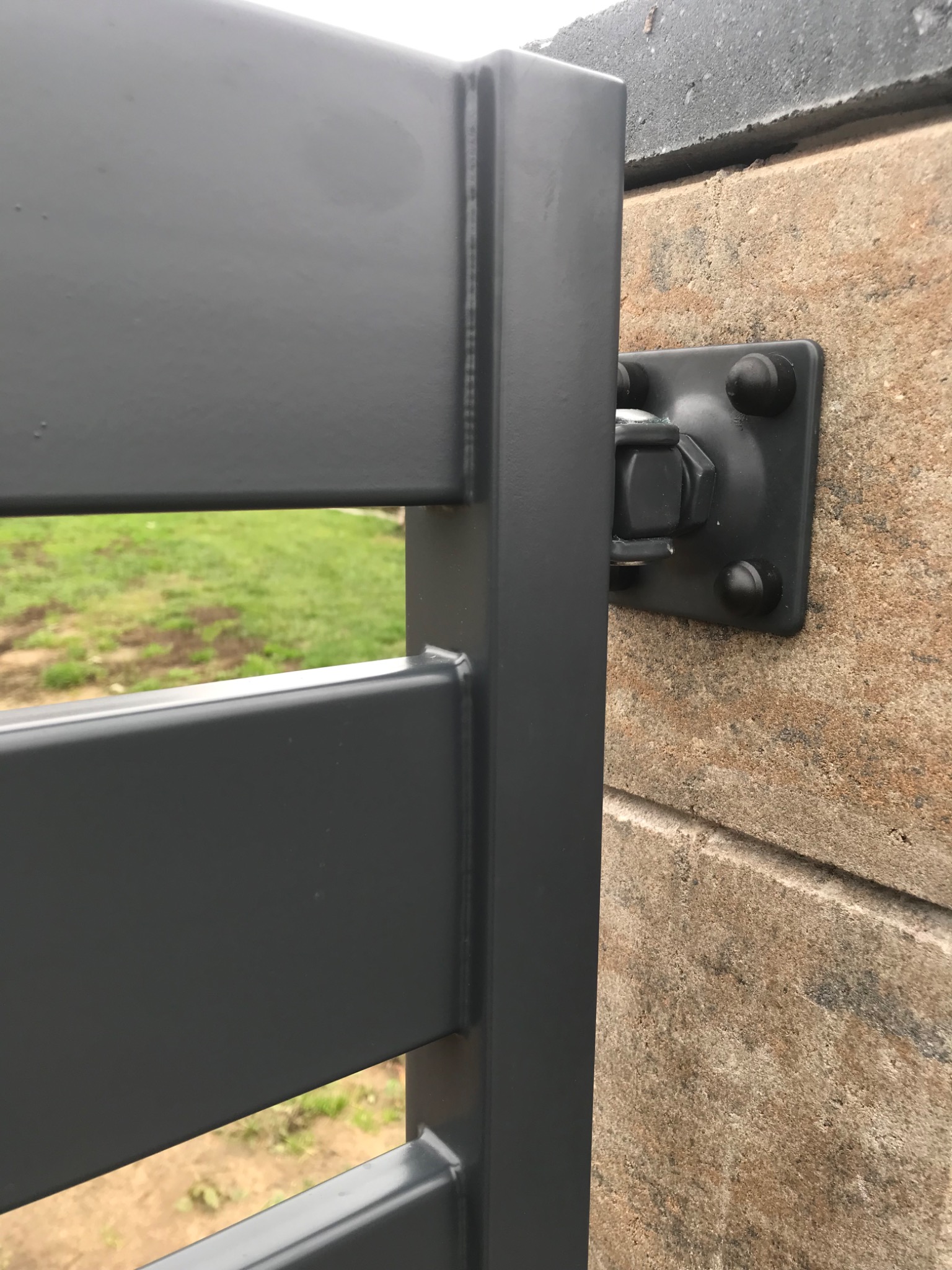 Szczegół mocowania metalowej bramy wjazdowej do murowanego słupka, widoczne śruby i spawane łączenia, trawa w tle