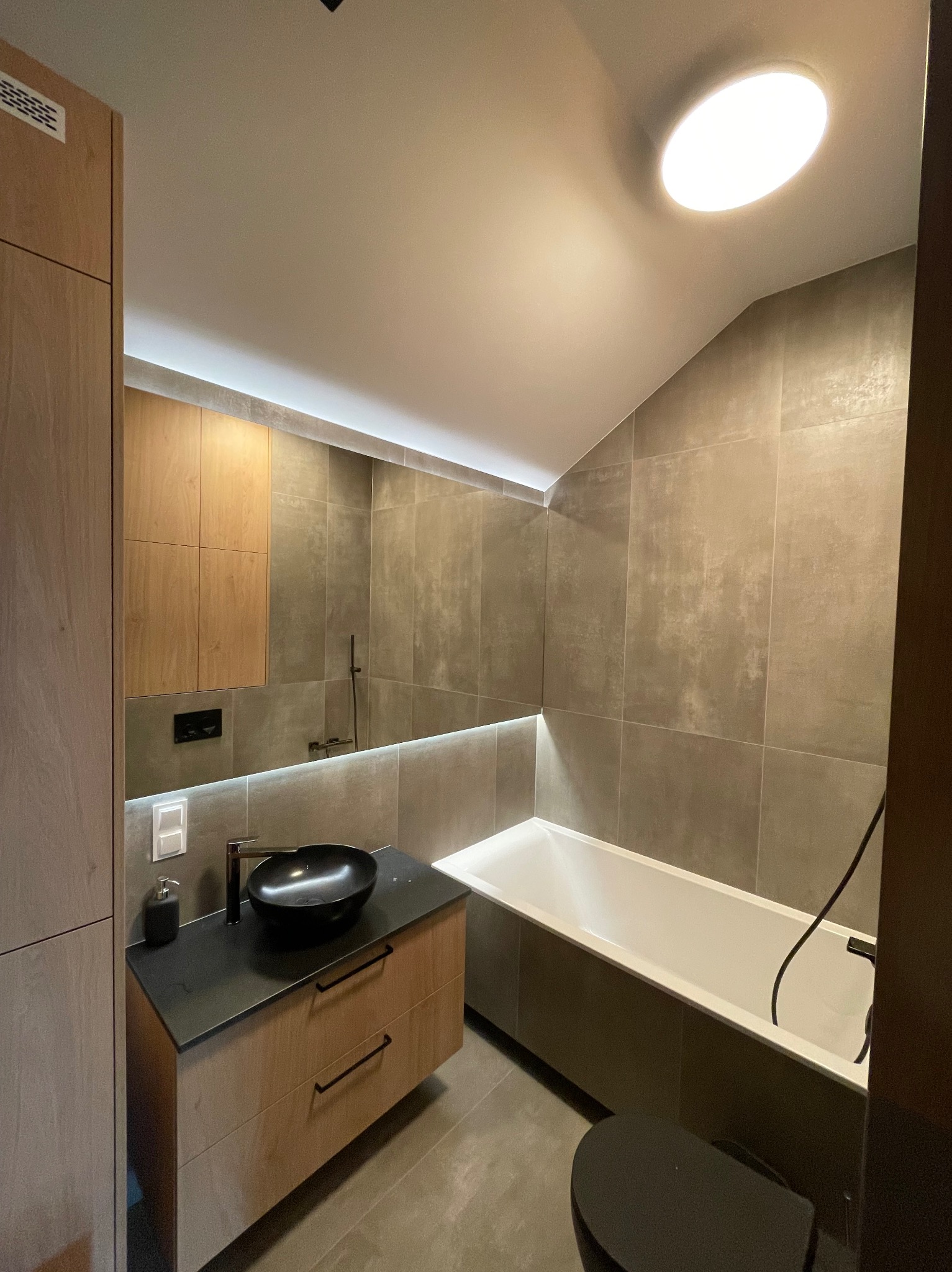 Nowoczesna łazienka na poddaszu z grafitowymi płytkami, drewnianą szafką z czarnym blatem i umywalką oraz podświetlaną wanną.