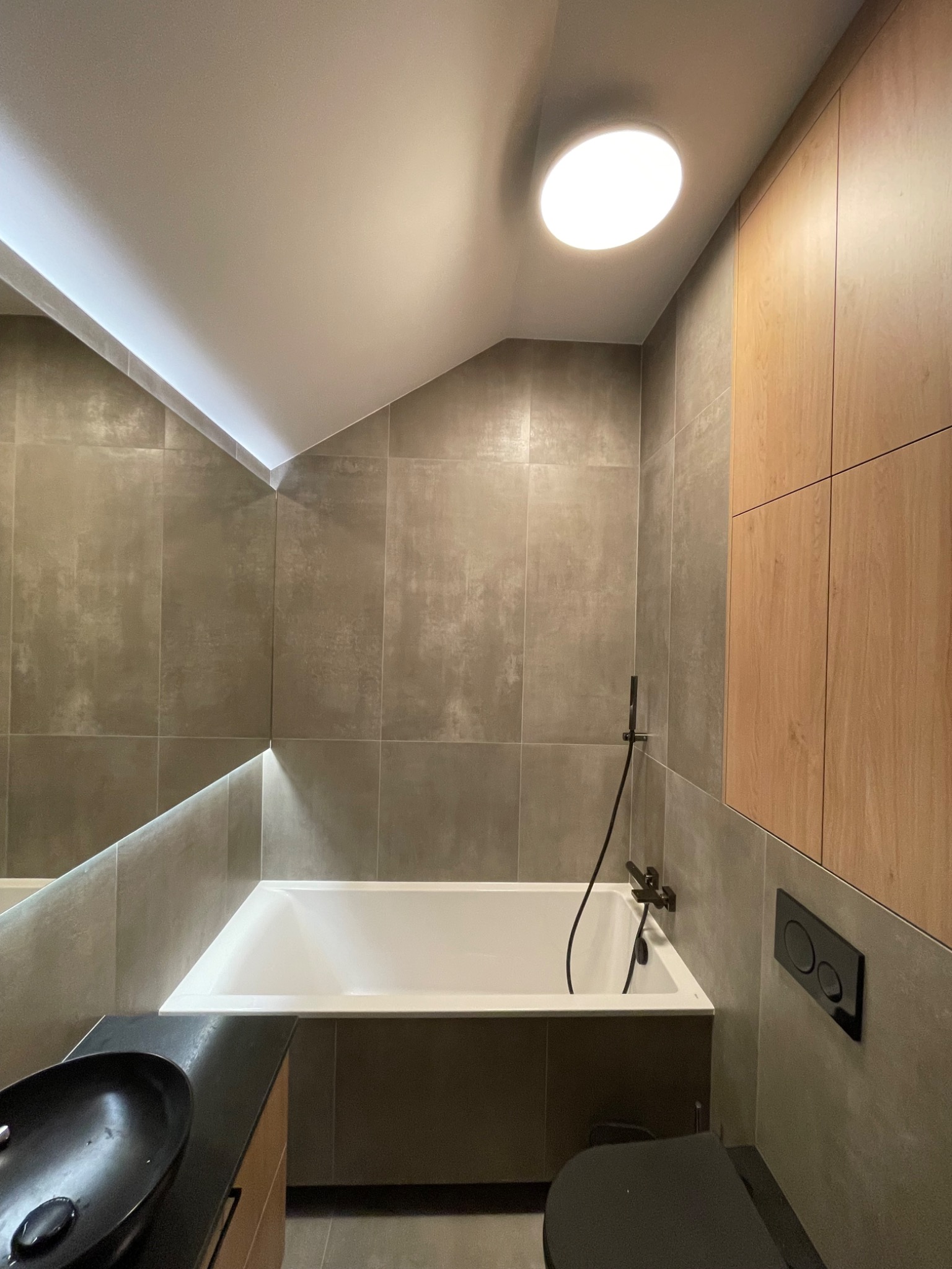 Nowoczesna łazienka z szarymi płytkami, białą wanną, czarną umywalką nablatową i drewnianymi szafkami, oświetlona okrągłą lampą sufitową i listwą LED pod skosem sufitu.
