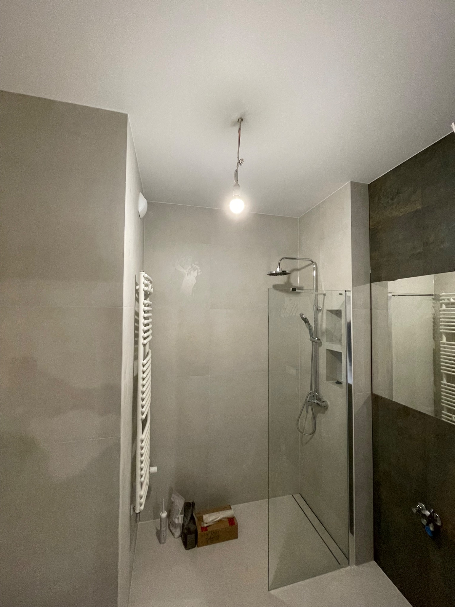 Nowoczesna kabina prysznicowa ze szklaną ścianą, chromowaną armaturą i grzejnikiem drabinkowym na ścianie z szarych płytek, widoczne materiały budowlane na podłodze, surowa żarówka zwisająca z sufitu.