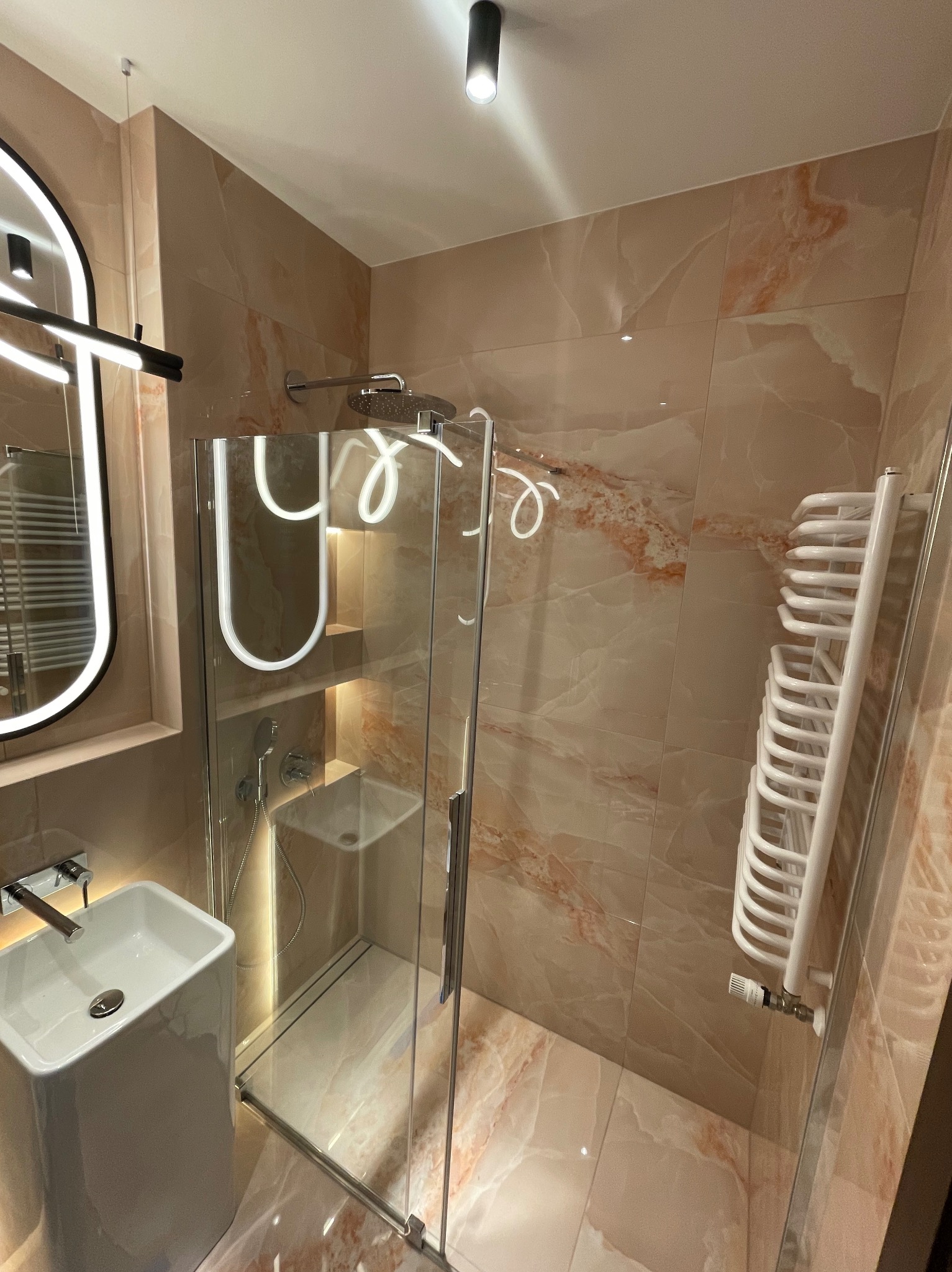 Nowoczesna łazienka z kabiną prysznicową typu walk-in, umywalką na postumencie i grzejnikiem drabinkowym na ścianie wyłożonej płytkami imitującymi marmur.