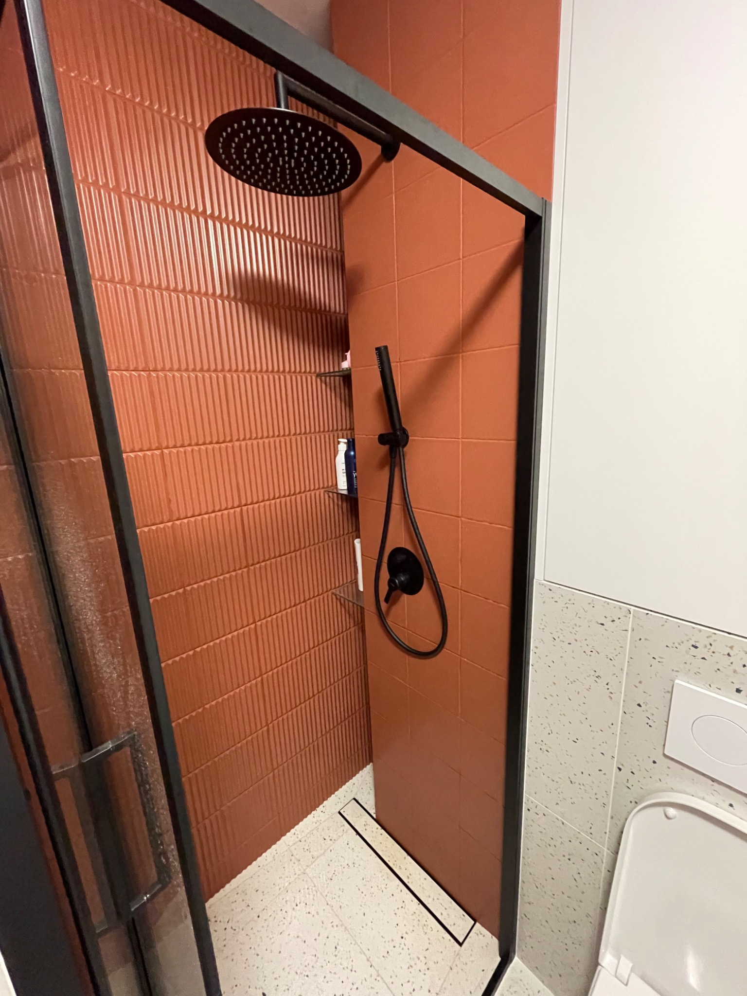 Nowoczesna kabina prysznicowa z czarną armaturą i terakotowymi płytkami o fakturze pionowych prążków, widok na deszczownicę i odpływ liniowy w posadzce wykończonej lastryko.