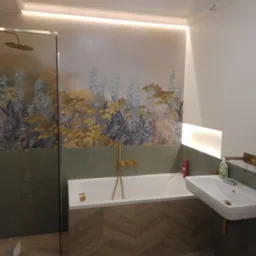 Nowoczesna łazienka z wanną, umywalką i kabiną prysznicową, wykończona płytkami w odcieniach zieleni i drewna, z dekoracyjną tapetą z motywem roślinnym i złotą armaturą. Na podłodze widoczne...