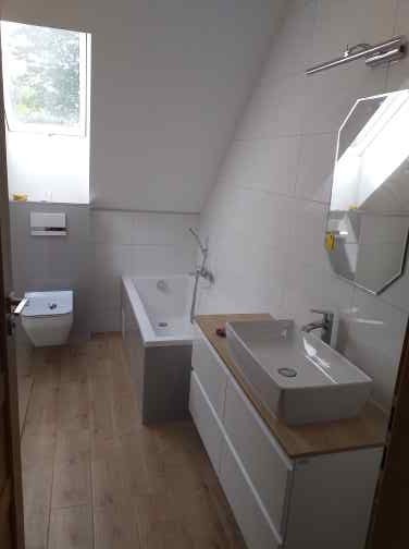 Jasna łazienka na poddaszu z drewnianą podłogą, białą wanną, umywalką na szafce z blatem drewnopodobnym i lustrem w geometrycznej ramie.