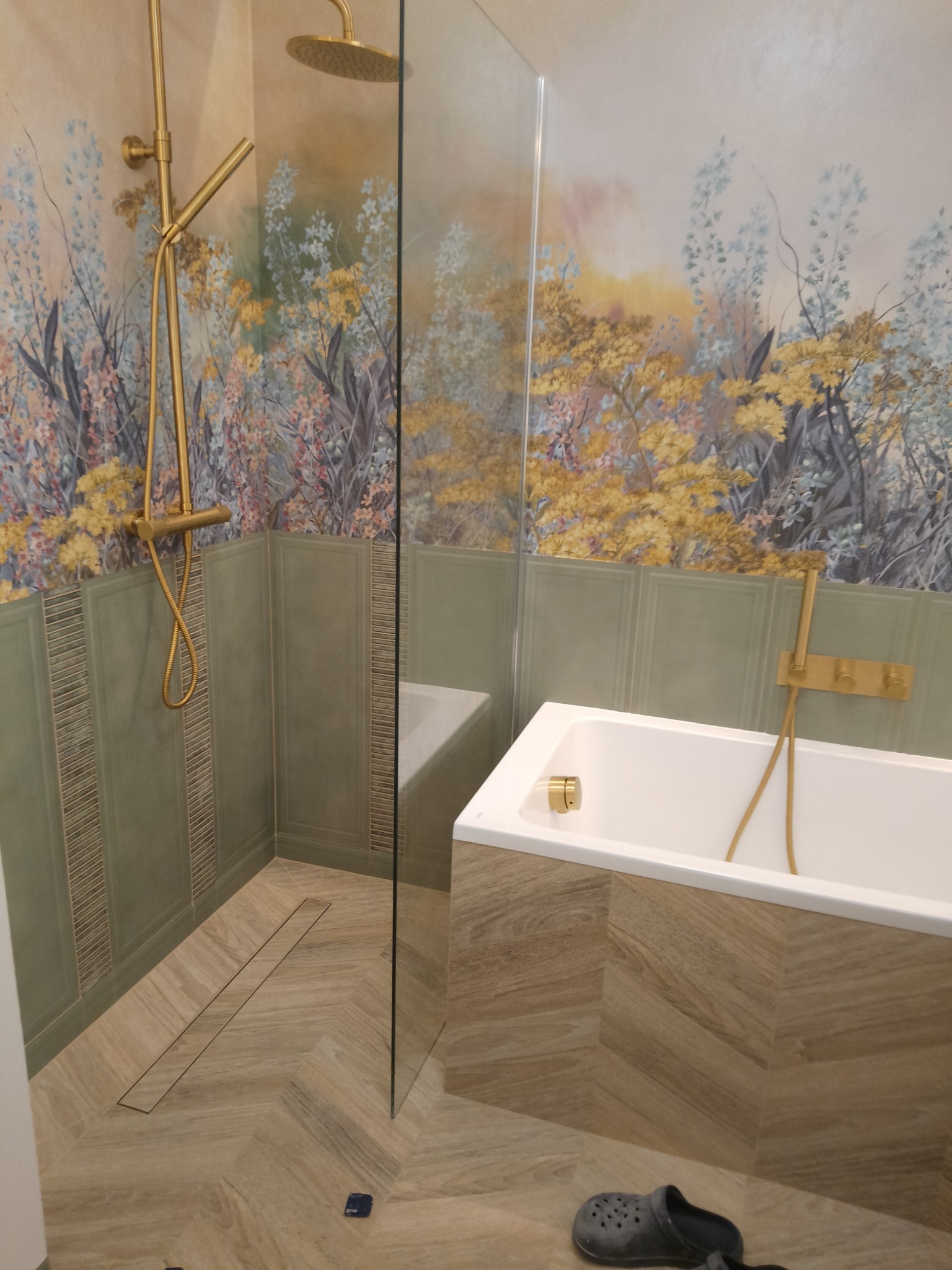 Nowoczesna łazienka z kabiną prysznicową typu walk-in, złotą armaturą, wanną obudowaną płytkami imitującymi drewno i dekoracyjną tapetą w kwiaty.