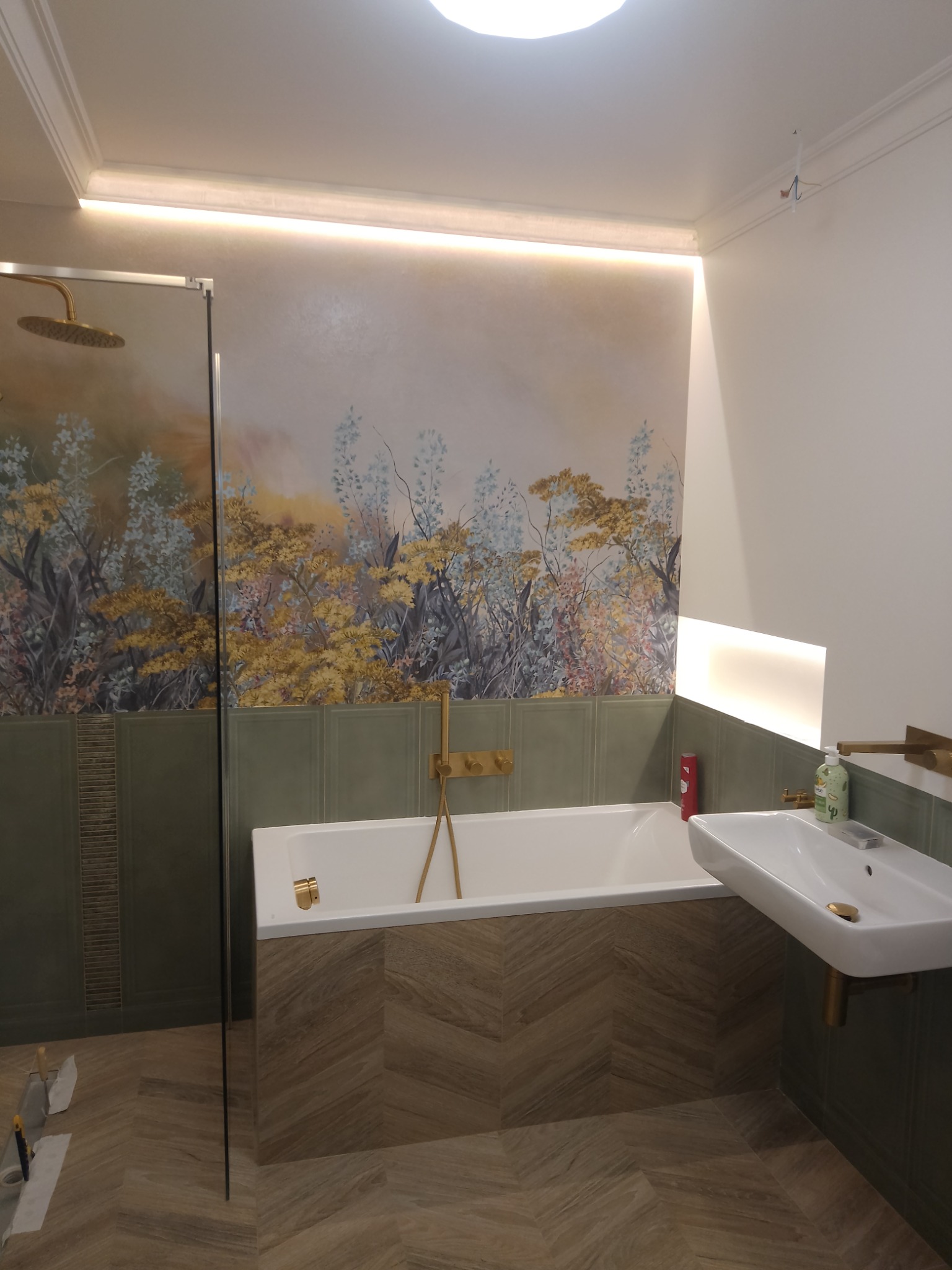 Nowoczesna łazienka z wanną, umywalką i kabiną prysznicową, wykończona płytkami w odcieniach zieleni i drewna, z dekoracyjną tapetą z motywem roślinnym i złotą armaturą. Na podłodze widoczne...