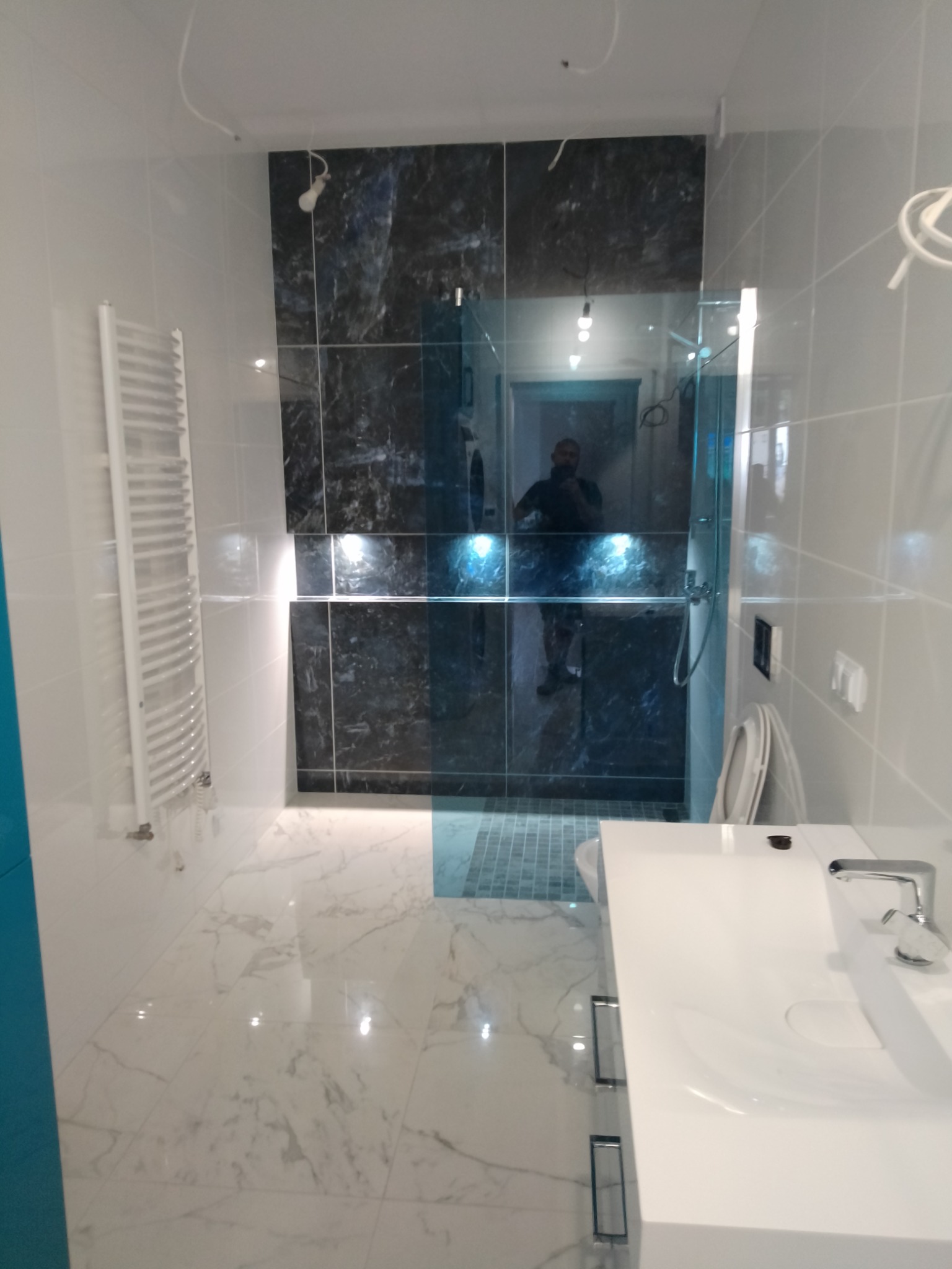 Nowoczesna łazienka z marmurową podłogą, kabiną prysznicową z ciemnymi płytkami imitującymi kamień, białym grzejnikiem drabinkowym i umywalką na szafce, widoczne niedokończone podłączenie...