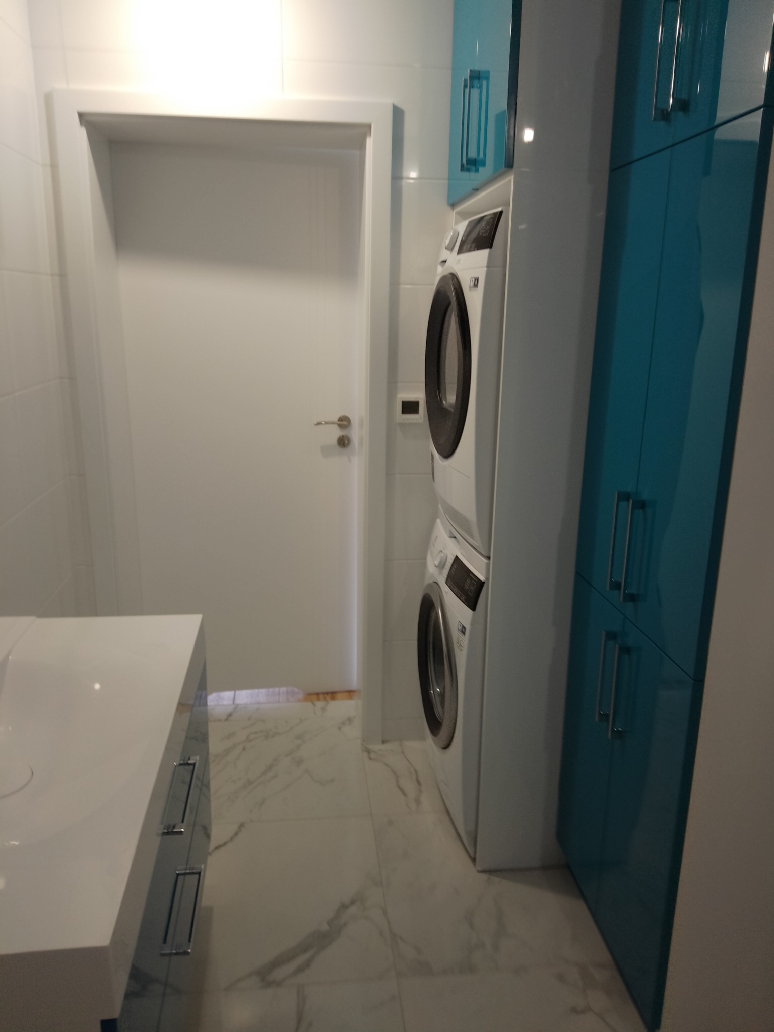 Nowoczesna łazienka z białą umywalką nablatową, dwiema pralkami w kolumnie i turkusowymi szafkami, wykończona białymi płytkami i marmurową podłogą.
