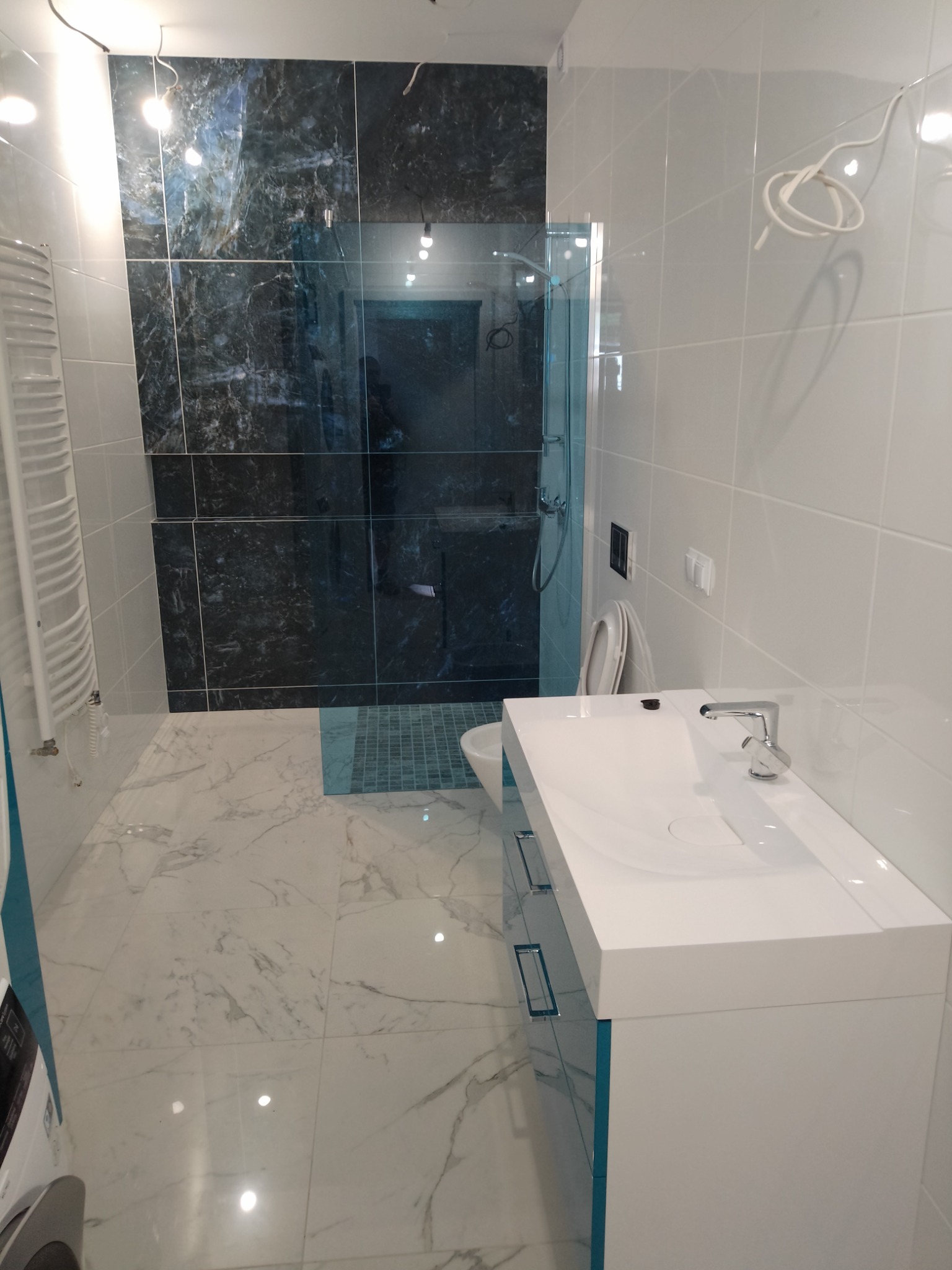 Nowoczesna łazienka z marmurową podłogą i ścianą prysznicową z ciemnego kamienia, biała umywalka z szafką i szklana kabina prysznicowa w tle.