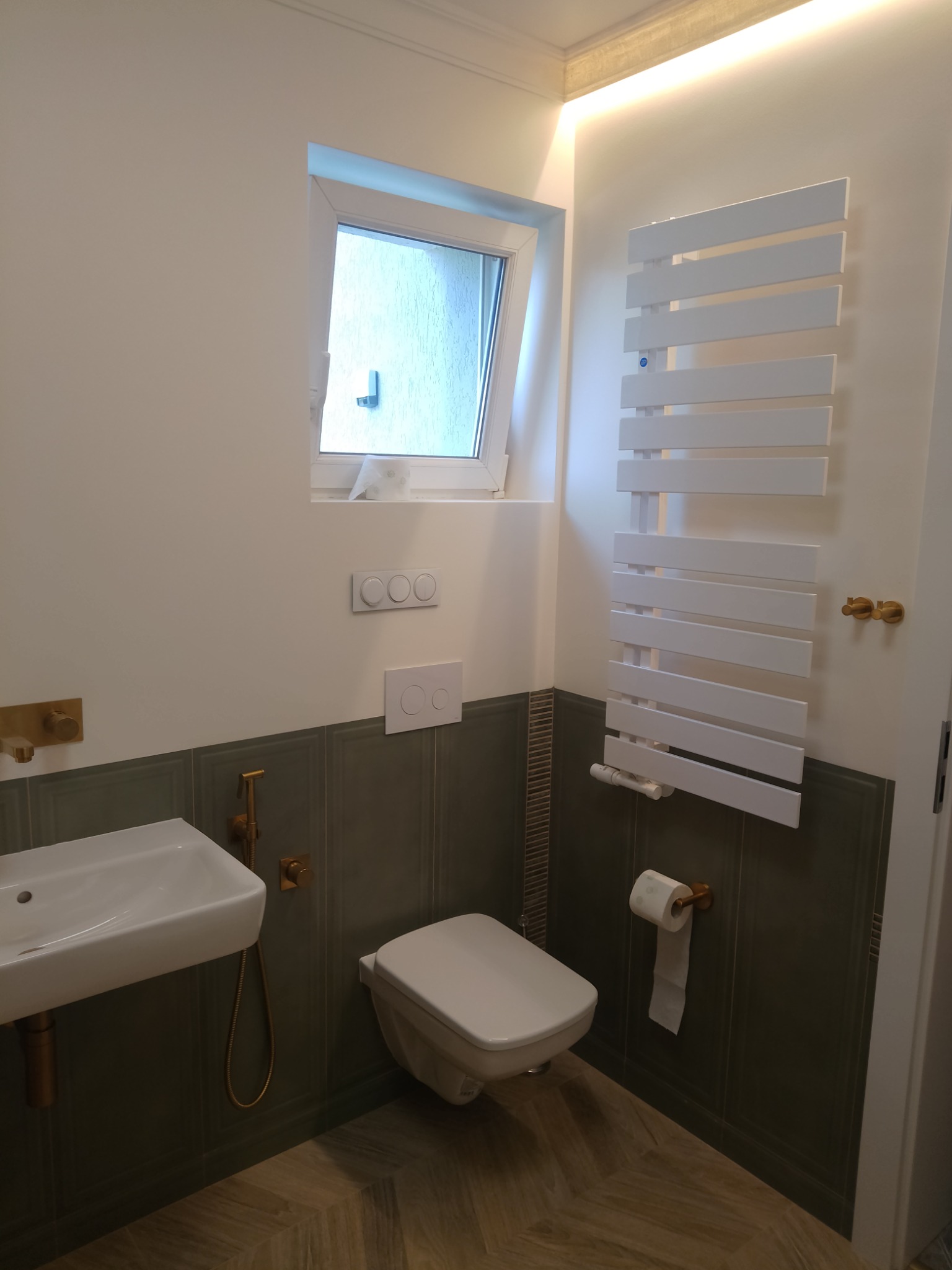 Nowoczesna łazienka z białą umywalką, toaletą podwieszaną i grzejnikiem drabinkowym, wykończona płytkami w odcieniach szarości i drewna, z akcentami w kolorze złotym.