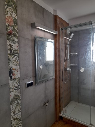 Nowoczesna łazienka z szarą płytką imitującą beton, dekoracyjnym pasem płytek z motywem roślinnym, podświetlanym lustrem i kabiną prysznicową z drewnianą obudową.