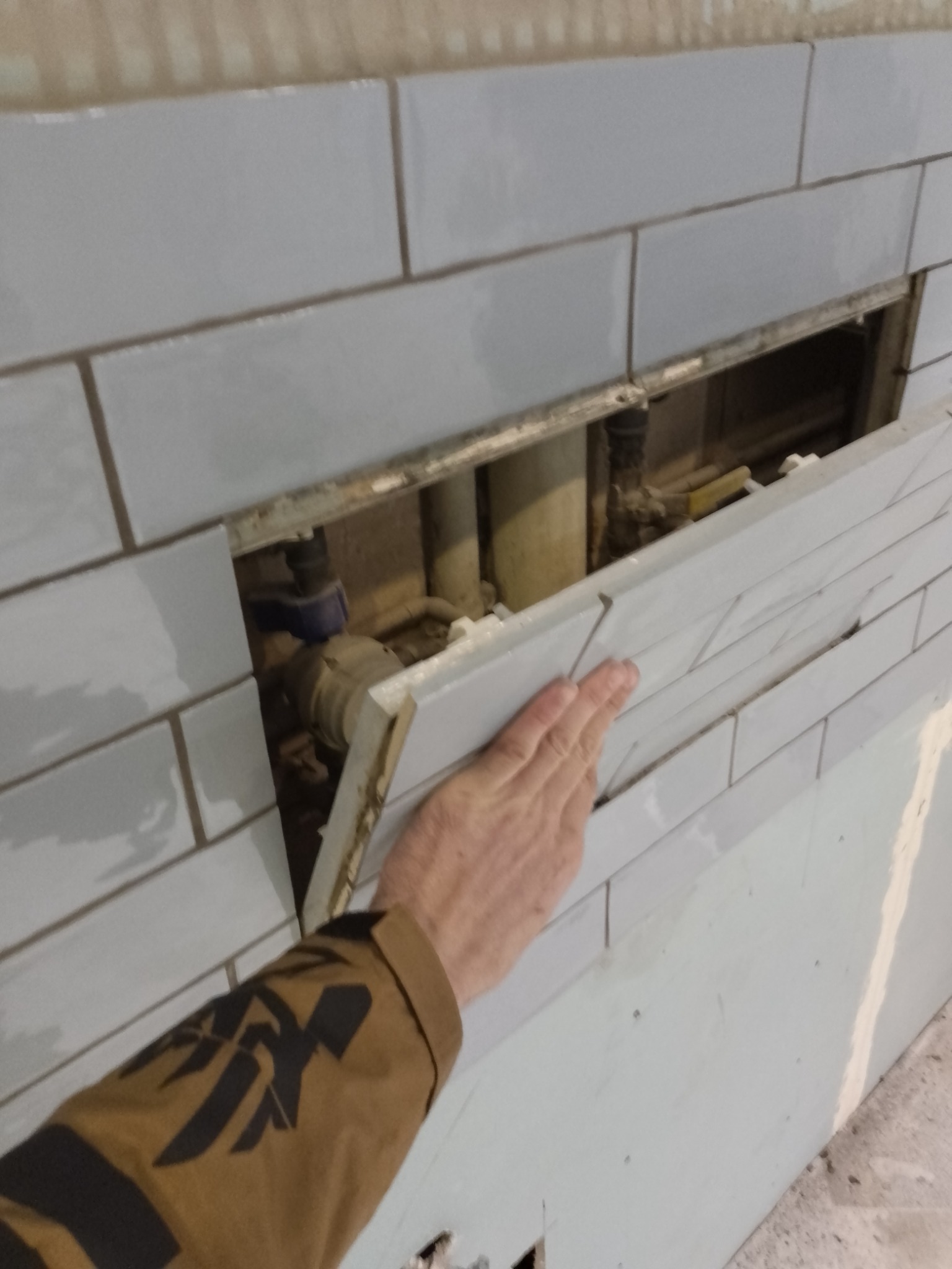 Układanie jasnoszarych płytek w łazience, widoczny fragment instalacji hydraulicznej za otworem rewizyjnym, ręka w brązowej kurtce z czarnym wzorem przytrzymuje płytkę.