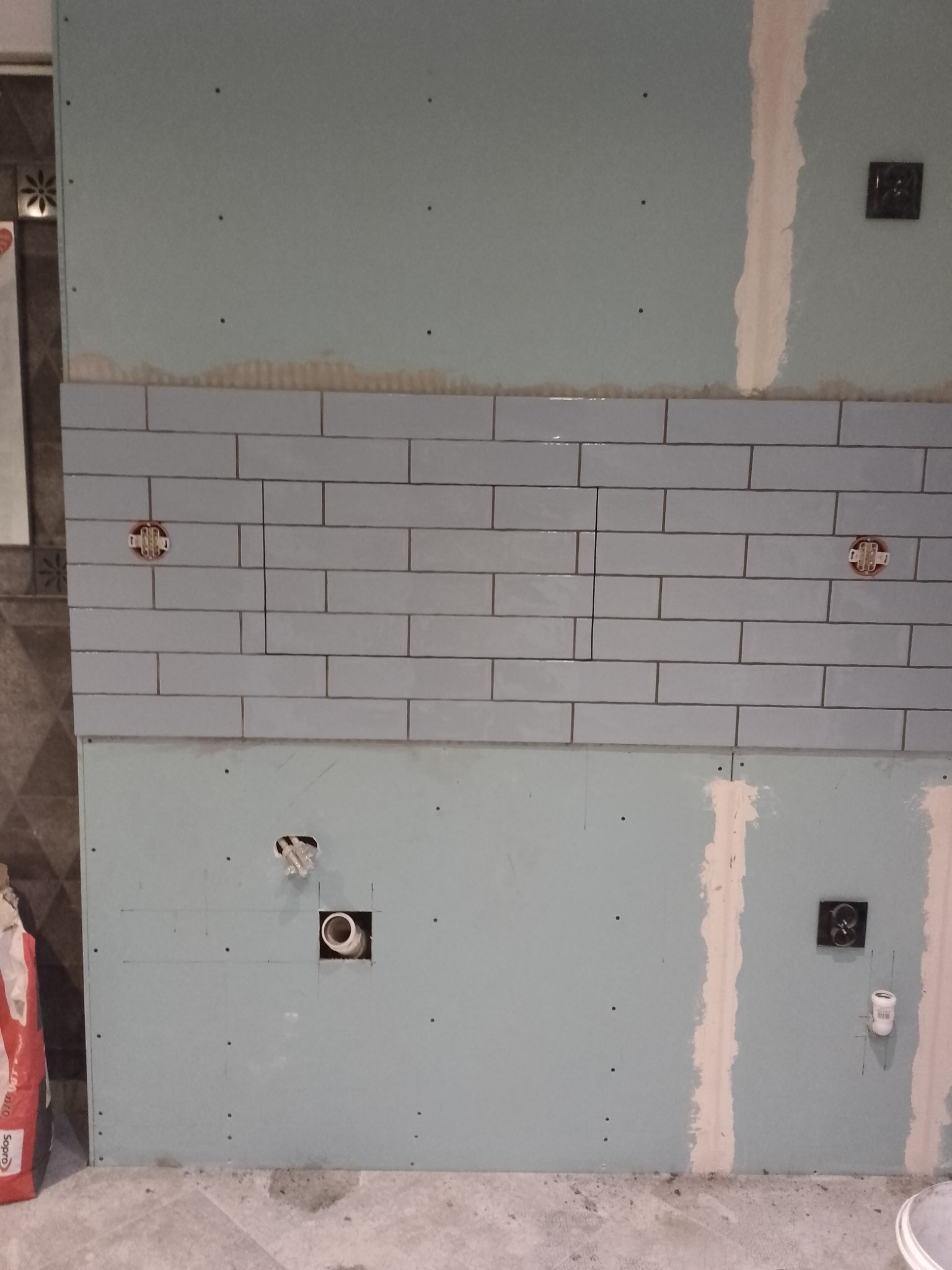 Ściana w trakcie wykańczania z częściowo położonymi, podłużnymi, jasnoniebieskimi płytkami ceramicznymi typu metro, widoczne puszki elektryczne i otwory na rury, płyta kartonowo-gipsowa oraz worek...