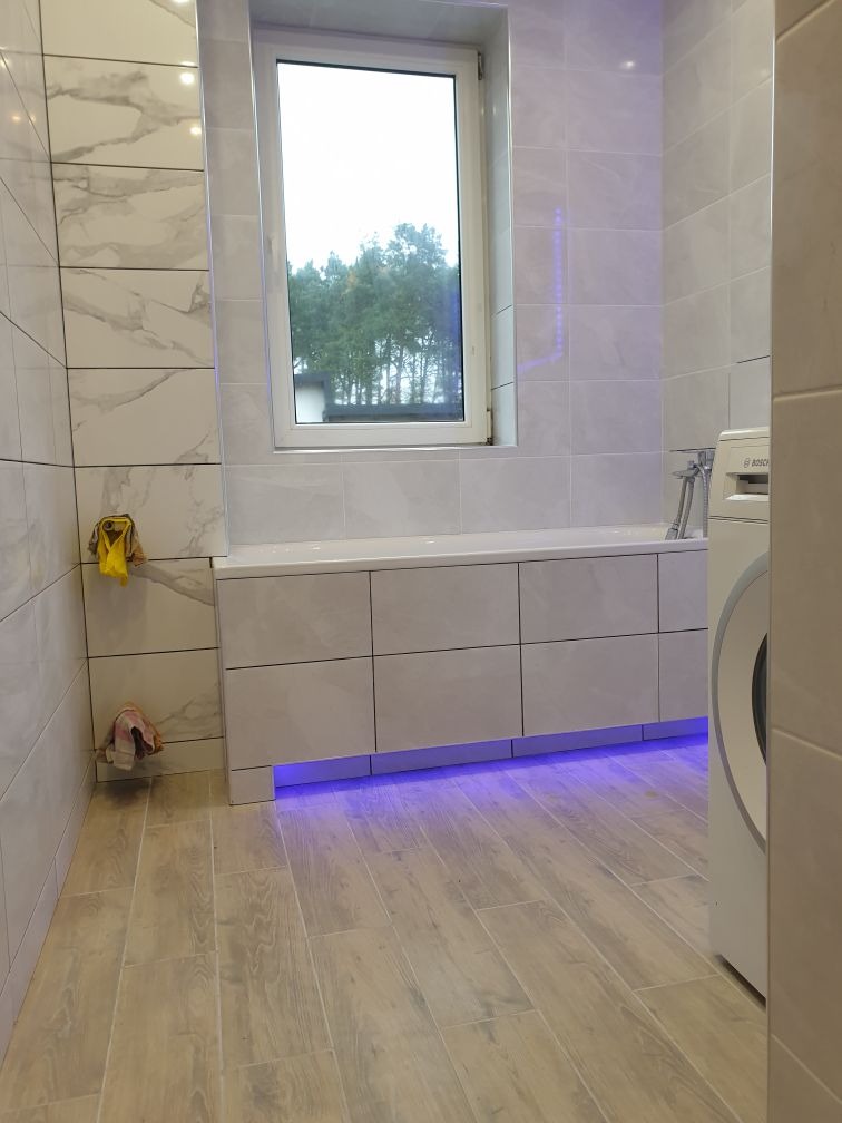Nowoczesna łazienka z białymi płytkami imitującymi marmur, podświetlaną szafką pod umywalką z niebieskim światłem LED i drewnopodobną podłogą, widok na okno z zielenią na zewnątrz.
