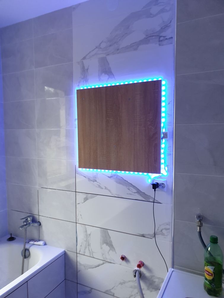 Wykończona łazienka z płytkami imitującymi marmur, drewnianą szafką z podświetleniem LED, widoczna bateria wannowa i butelka płynu do czyszczenia.