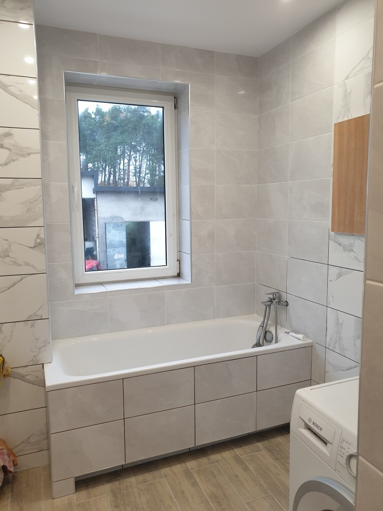 Wykończona łazienka z wanną obudowaną płytkami imitującymi marmur, jasnoszare płytki na ścianach, okno z widokiem na drzewa i pralką Bosch w rogu.