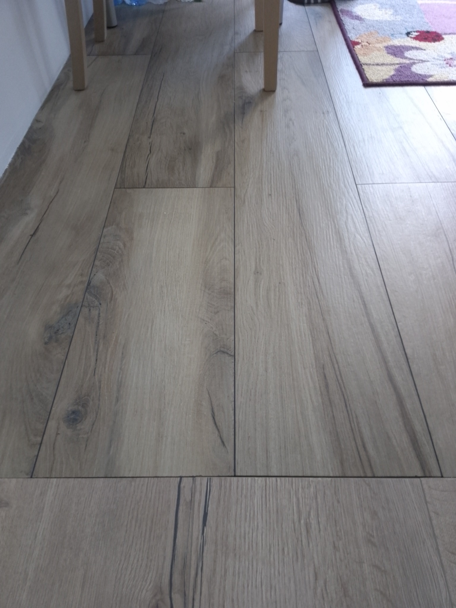 Ułożona podłoga z płytek imitujących drewno, widoczne naturalne słoje i sęki, fragment dywanu z kwiatowym wzorem w rogu.