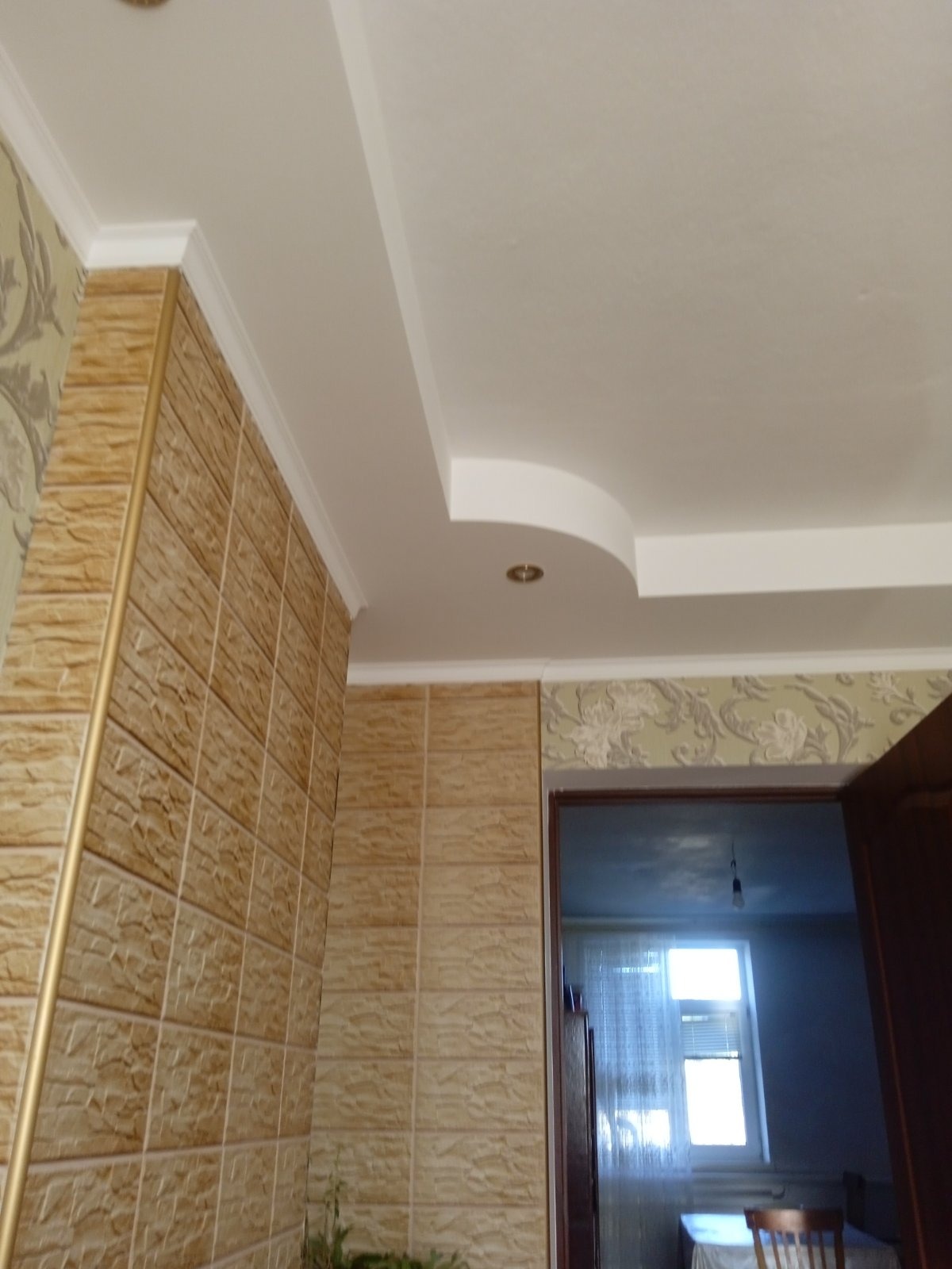 Fragment wnętrza z dekoracyjną ścianą z paneli imitujących kamień, białym sufitem z podwieszanym elementem i oświetleniem punktowym, widok na pokój przez otwarte drzwi.