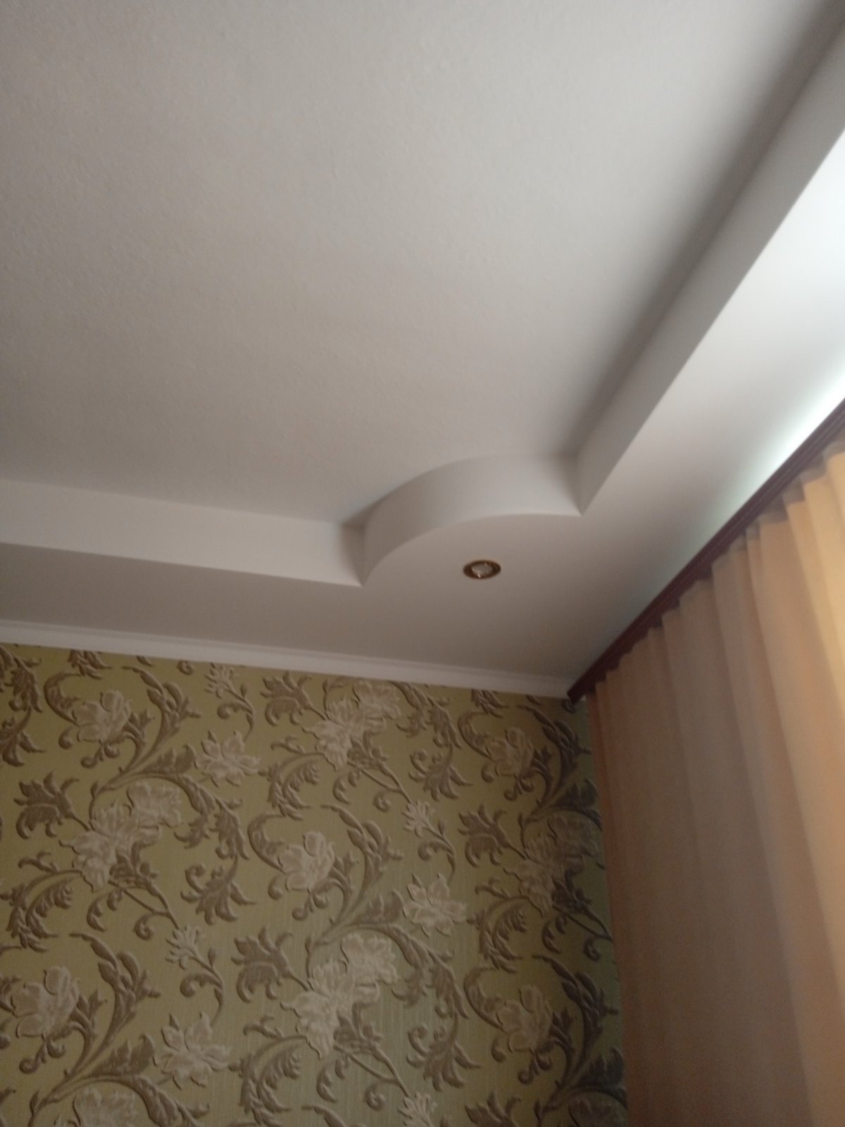 Biały, wielopoziomowy sufit z dekoracyjnym oświetleniem punktowym, kontrastujący z wzorzystą tapetą w odcieniach beżu i brązu oraz zasłoną w rogu pomieszczenia.