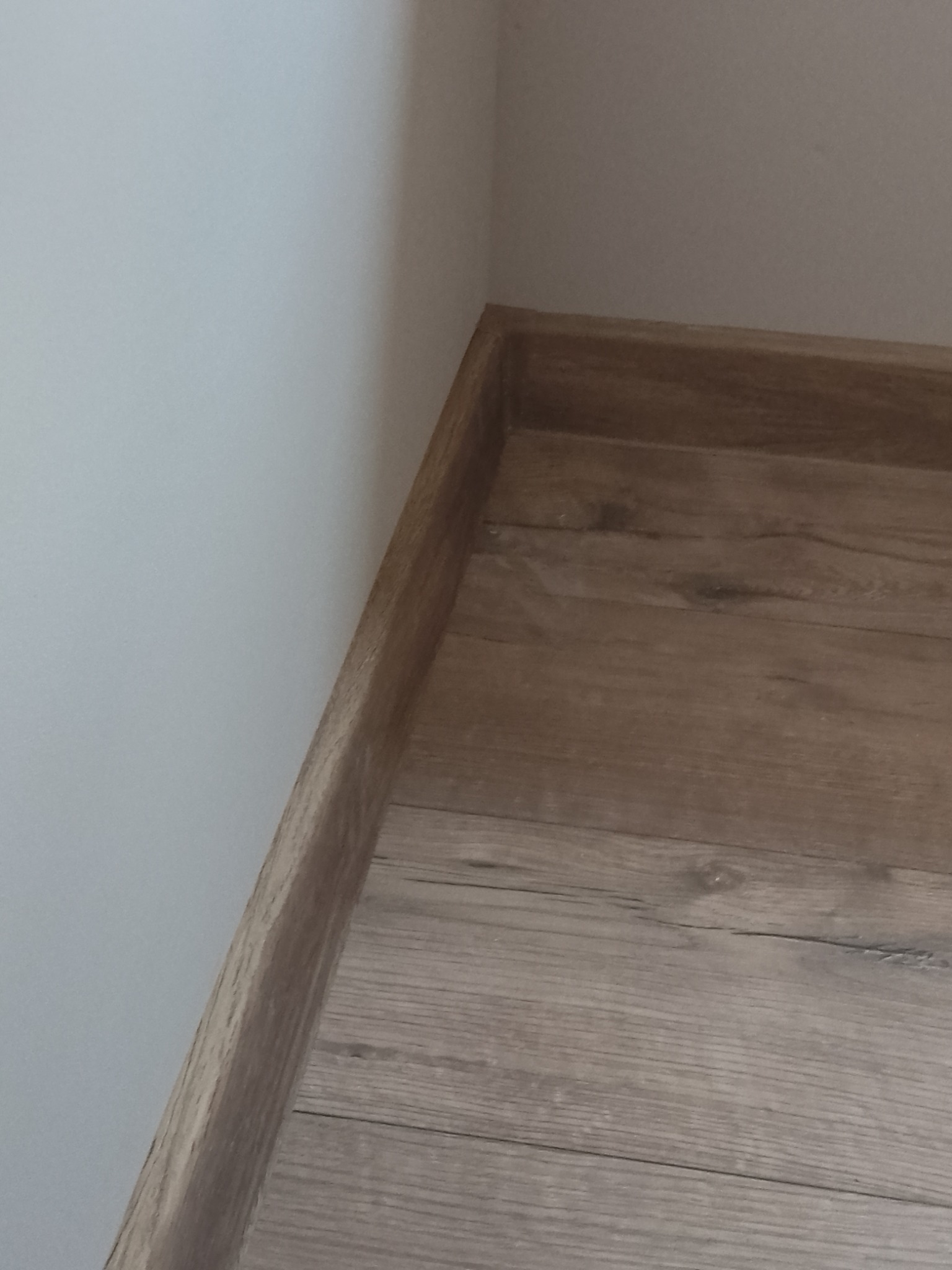 Szczegółowe ujęcie narożnika pokoju po remoncie: połączenie jasnej ściany i podłogi z paneli imitujących drewno, wykończonych listwą przypodłogową w odcieniu zbliżonym do podłogi.