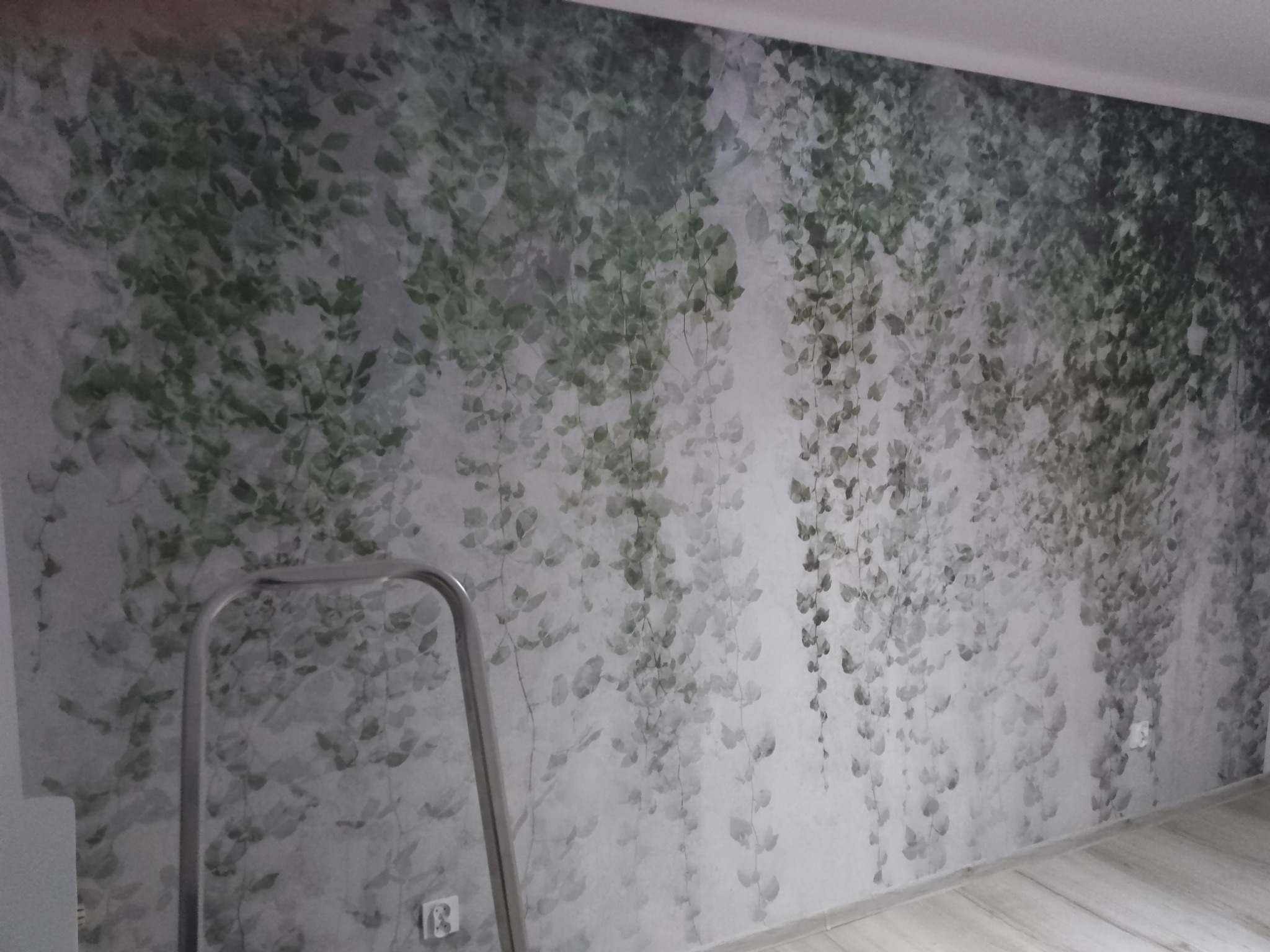 Ściana pokryta fototapetą z motywem pnących roślin, widoczna drabina malarska, fragment podłogi z paneli.