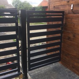 GKDSpaw - Nowoczesna, pozioma brama wjazdowa z czarnych, metalowych profili, zamontowana przy drewnianym ogrodzeniu, widok z bliska na zamek i zawiasy.