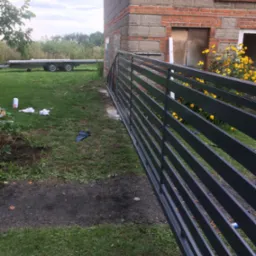 Nowoczesna, pozioma brama wjazdowa z metalowych profili, świeżo pomalowana na ciemnoszary kolor, ustawiona przy ceglanym budynku w trakcie budowy, z widocznym przyczepą i polnymi kwiatami w tle.