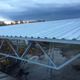 Konstrukcja metalowa z dachem z blachy trapezowej w trakcie montażu, widoczne elementy konstrukcyjne, drabina i sprzęt rolniczy w tle, pochmurne niebo.