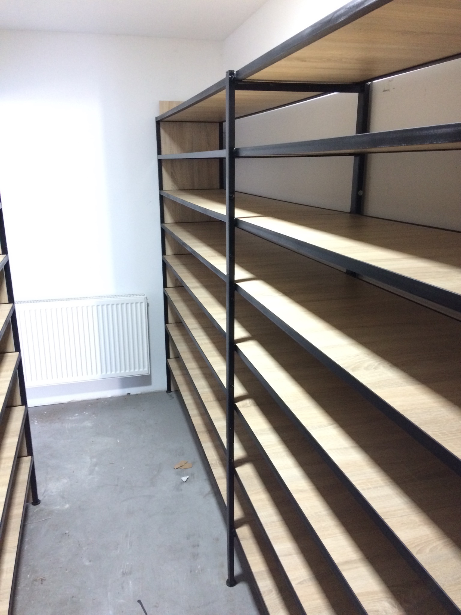 Metalowy regał z drewnianymi półkami, ustawiony w rogu pomieszczenia z betonową podłogą i grzejnikiem na ścianie.