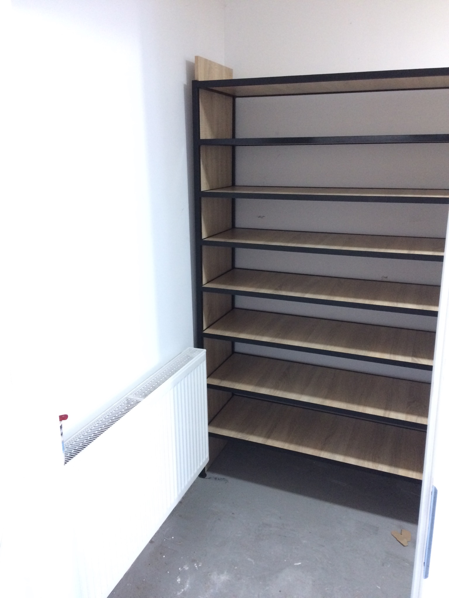 Metalowy regał z drewnianymi półkami, czarne ramy, osiem półek, zamontowany w rogu pokoju obok białego grzejnika.