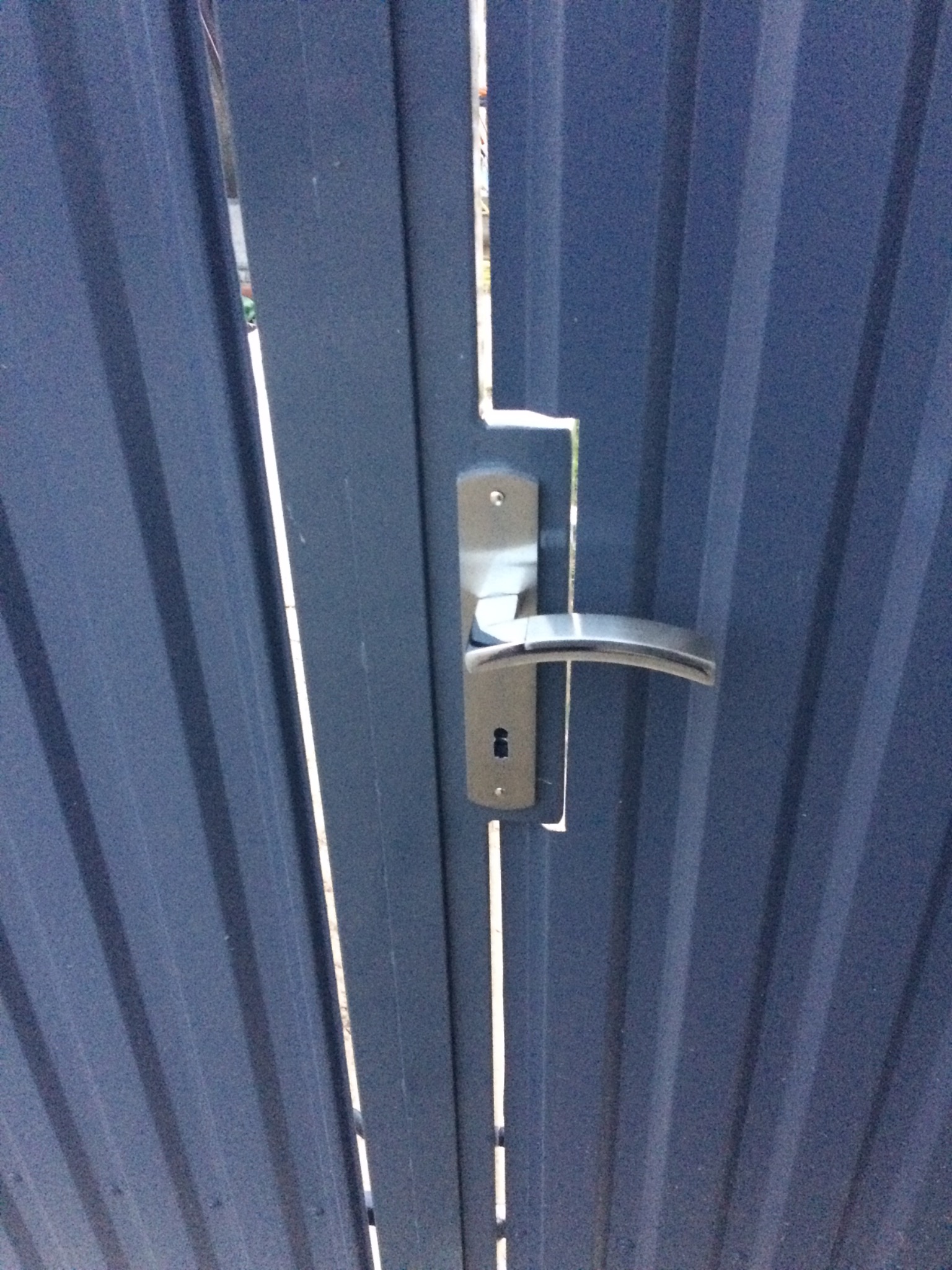 Fragment metalowej bramy wjazdowej z zamontowaną srebrną klamką i widocznym zamkiem, pionowe żłobienia na powierzchni bramy.