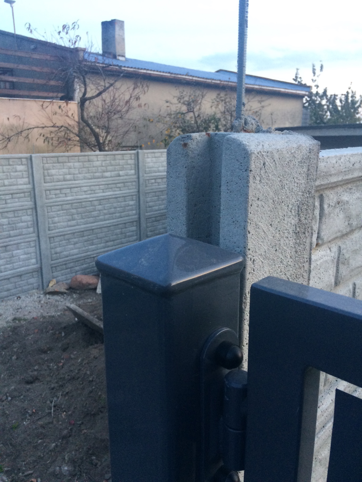 Fragment nowej bramy wjazdowej z czarnymi elementami metalowymi i betonowym słupkiem w trakcie montażu, widoczne śruby mocujące i betonowy płot w tle.