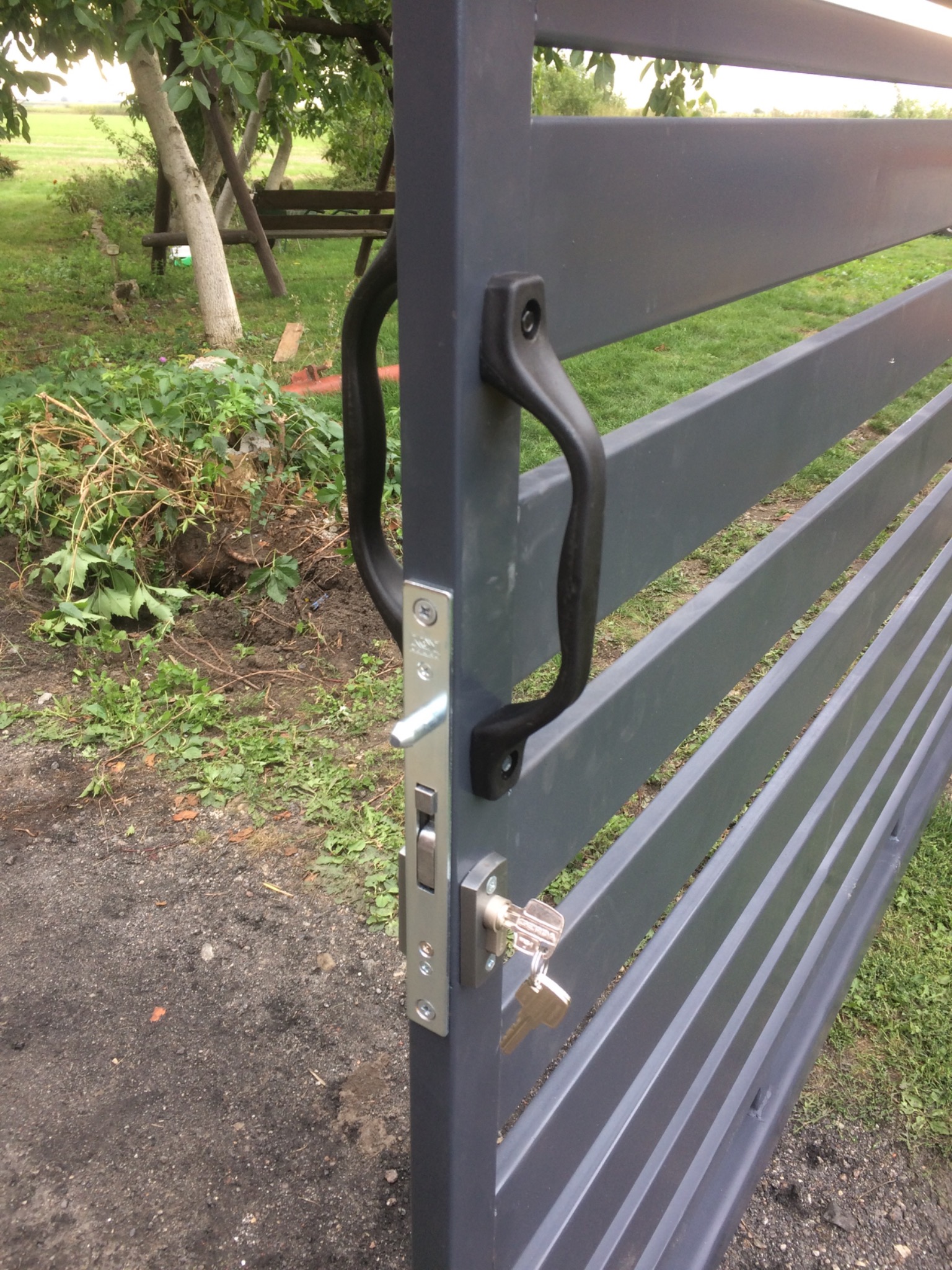 Szara, pozioma brama wjazdowa z czarną klamką i zamkiem z kluczem, na tle zielonego trawnika i drzew