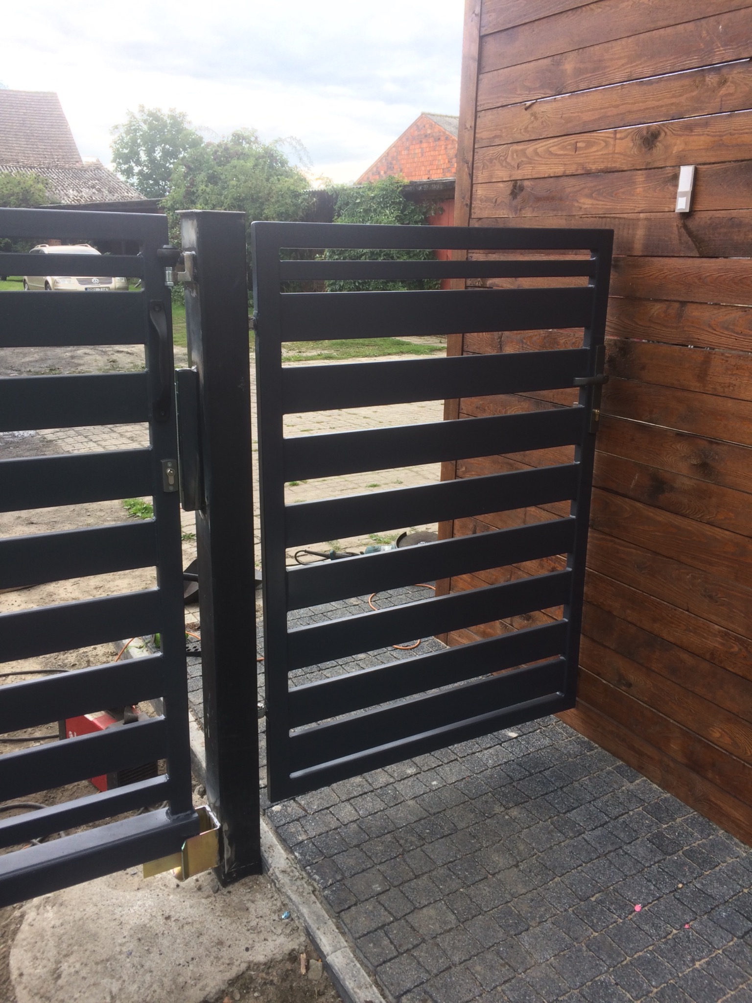 Nowoczesna, pozioma brama wjazdowa z czarnych, metalowych profili, zamontowana przy drewnianym ogrodzeniu, widok z bliska na zamek i zawiasy.