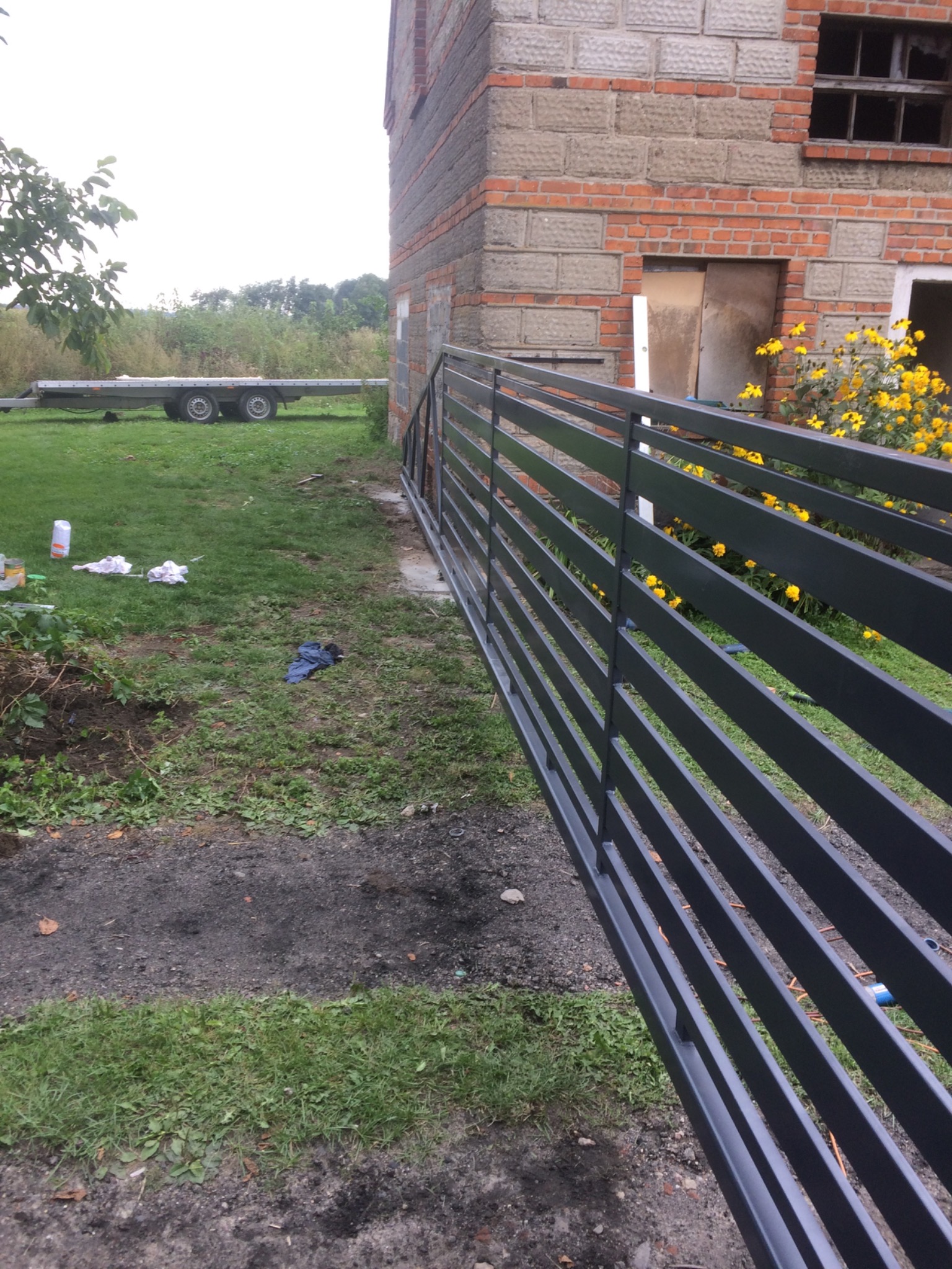 Nowoczesna, pozioma brama wjazdowa z metalowych profili, świeżo pomalowana na szaro, prowadząca do budynku z cegły, na trawniku widoczne ślady prac i narzędzia.